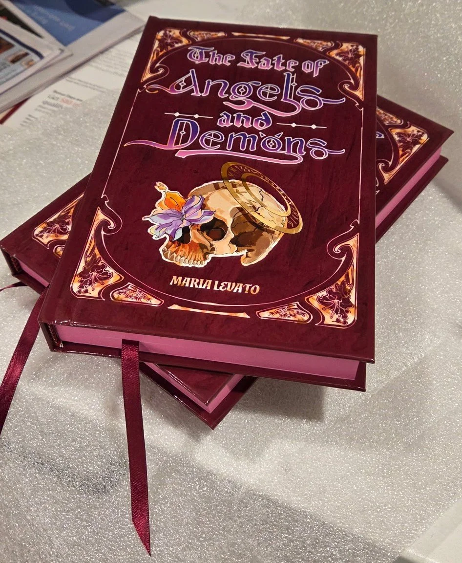 A hardcover book titled "The Fade of Angels and Demons" by Maria Levato with a dark red cover, decorated with gold and purple accents, featuring a skull with a purple flower and a halo on its cover, resting on a glittery white surface.