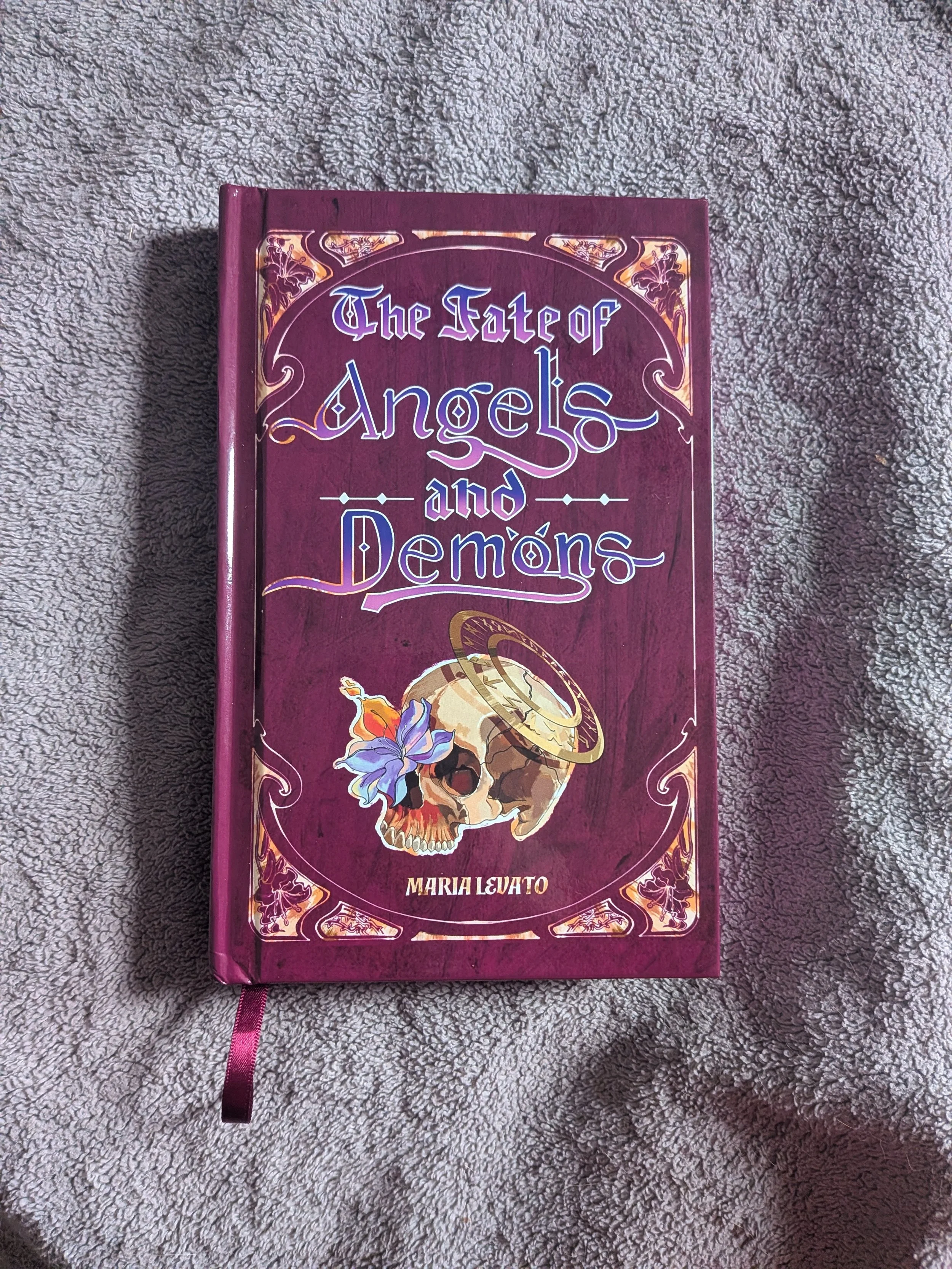 The Fate of Angels and Demons by Maria Levato (special edition hardcover). It shows a burgandy textured background with the title "The Fate of Angels and Demons" written in purple/pink ombre letters. Cover has skull w/ fire and a runic halo.