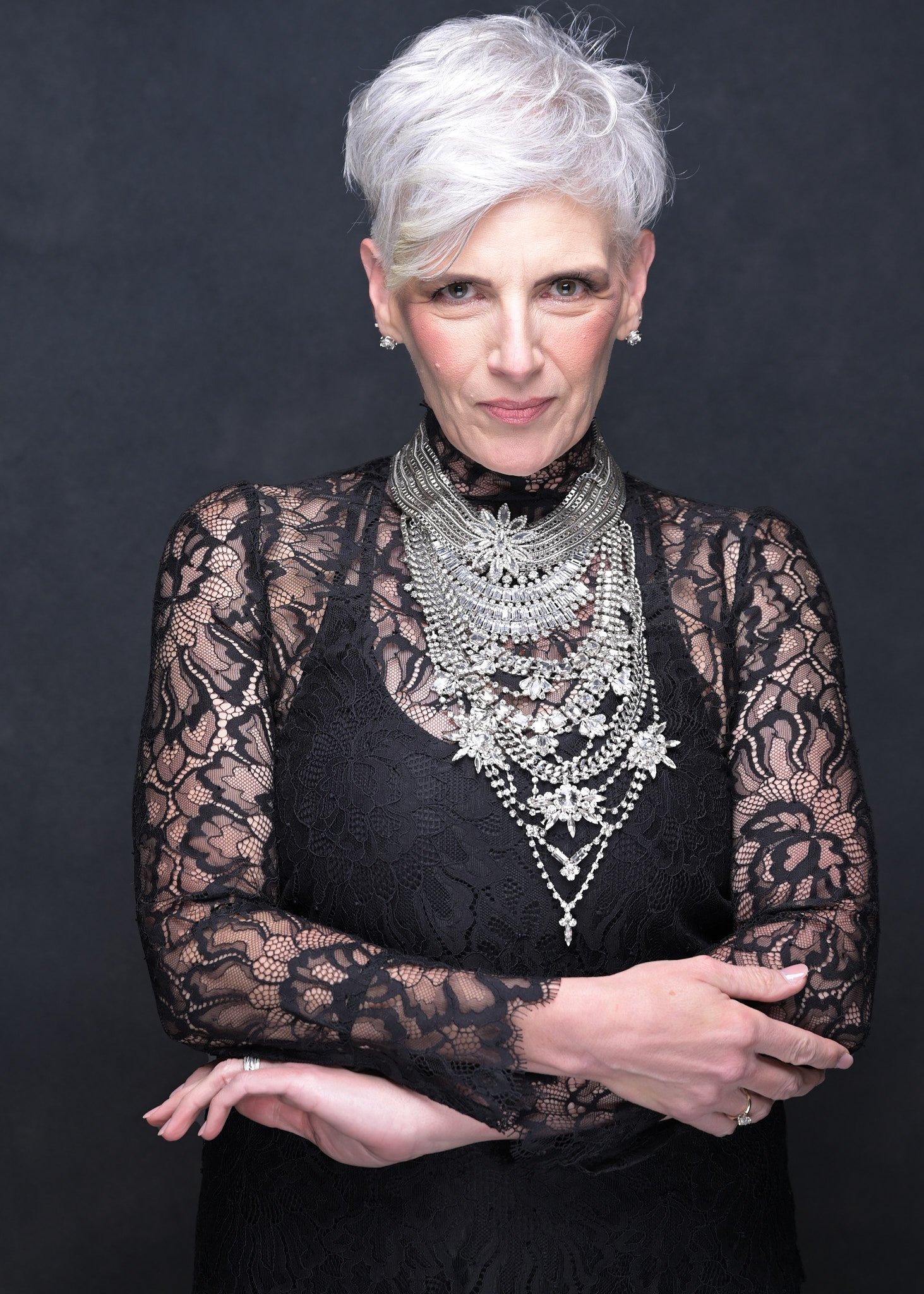A woman with short white hair wearing a black lace top and multiple layered silver necklaces standing against a dark background.  Empowering women, headshots and branding, age campaign