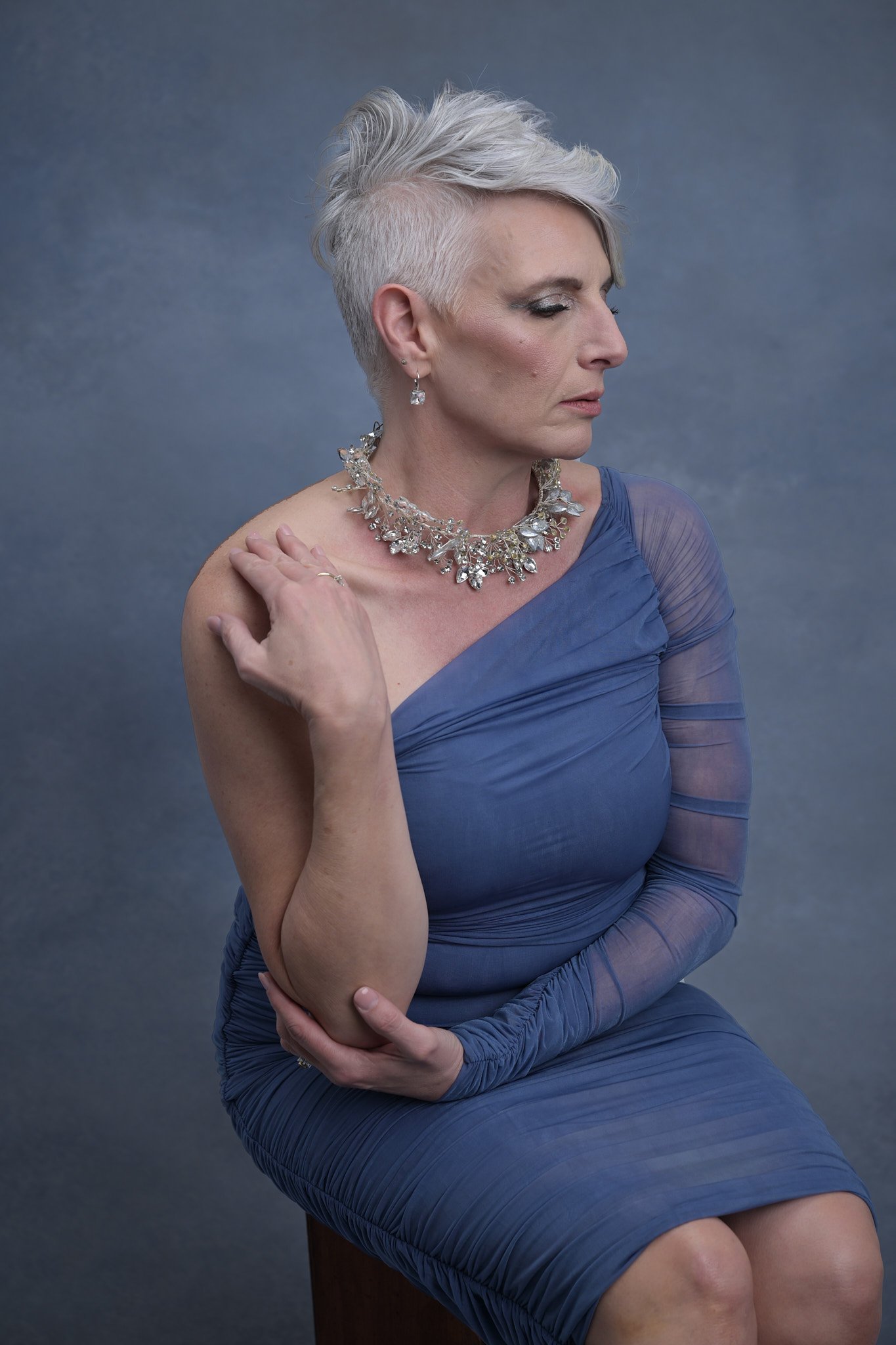 A woman with short, styled gray hair, wearing a one-shoulder blue dress with sheer sleeve detail, adorned with a large, elaborate diamond necklace and earrings, seated with a thoughtful expression against a gray background.   Empowering women, headsh