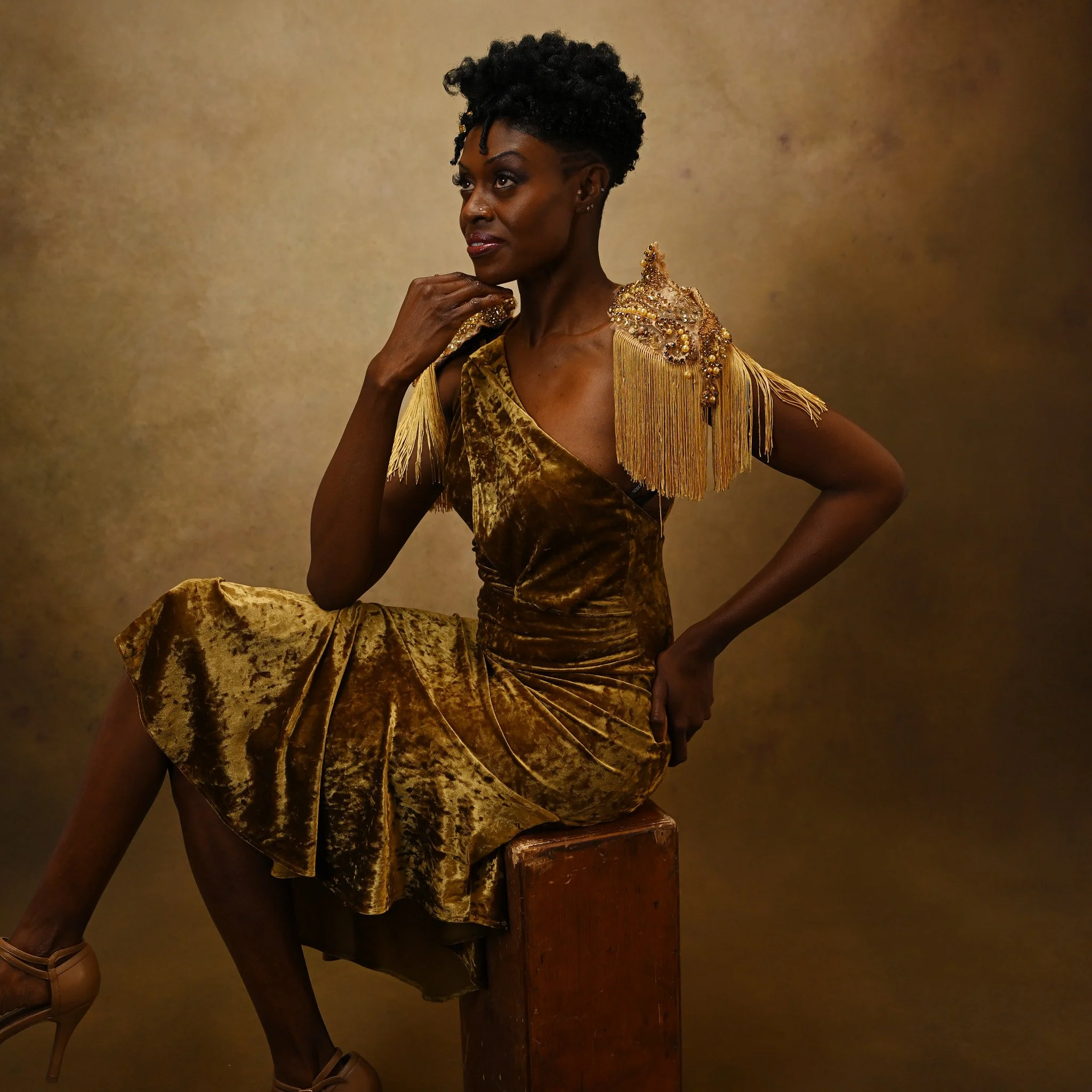 A woman wearing a gold velvet dress with ornate shoulder embellishments seated on a wooden block against a textured brown background.   Empowering women, headshots and branding, age campaign