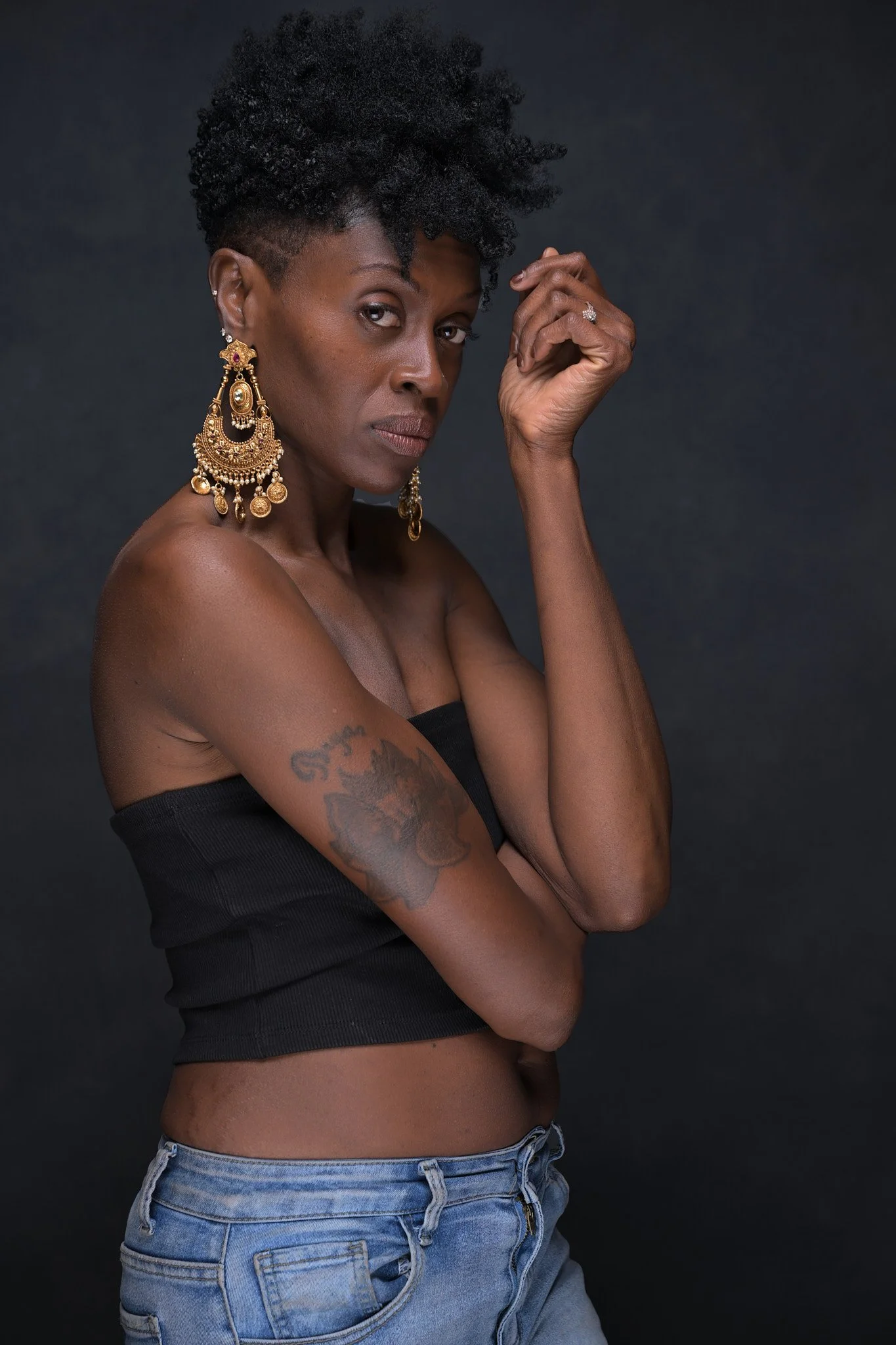A woman with short, curly black hair and dark skin is wearing large gold earrings, a black strapless top, and blue jeans. She has a tattoo on her upper arm and is looking directly at the camera with a serious expression, posing against a dark backgro