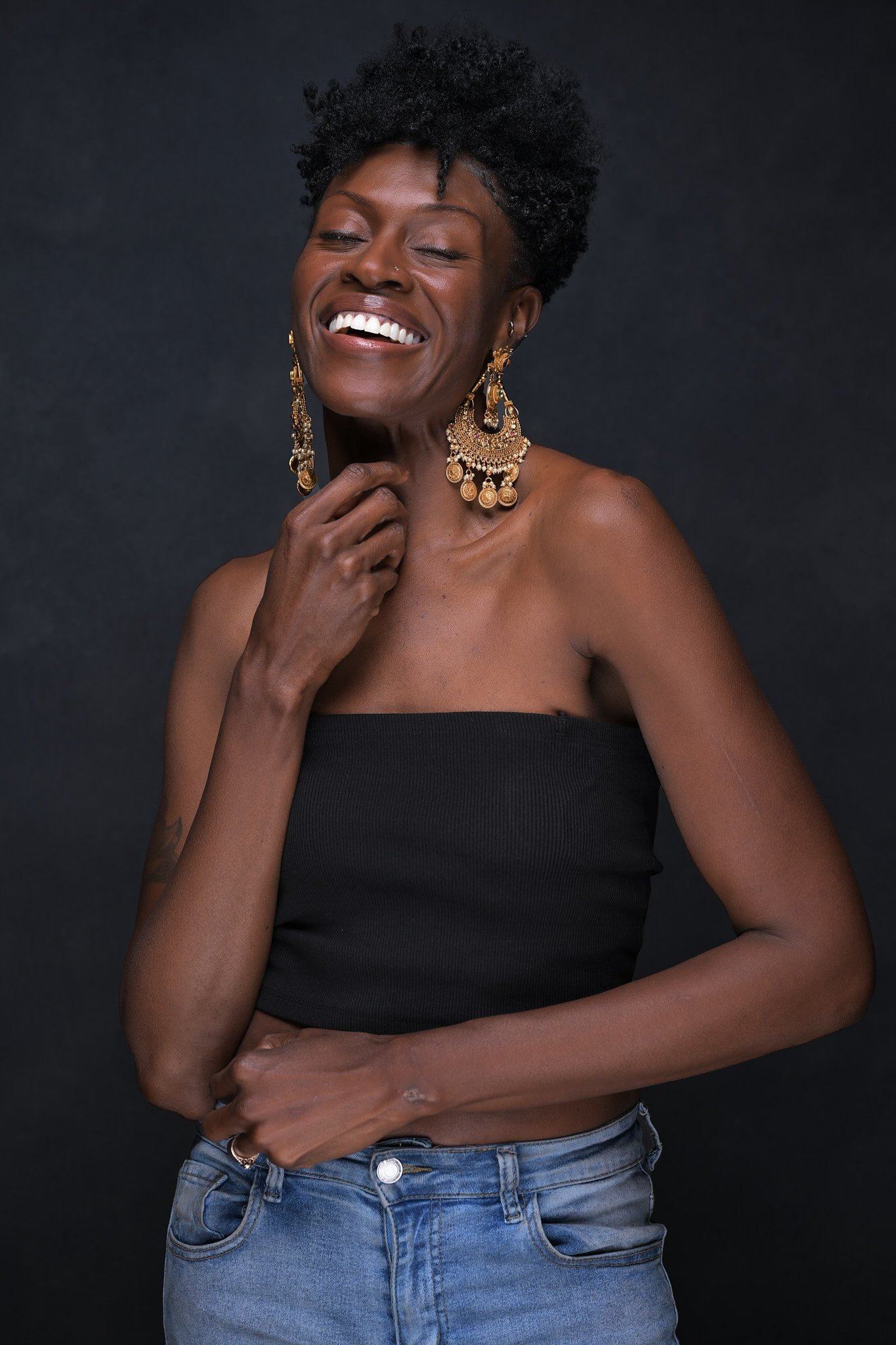 Portrait of a woman with short, curly black hair and dark skin, smiling with eyes closed, wearing large gold earrings, a black strapless top, and blue jeans against a dark background. Empowering women, headshots, and Branding sessions.  Age campaign.