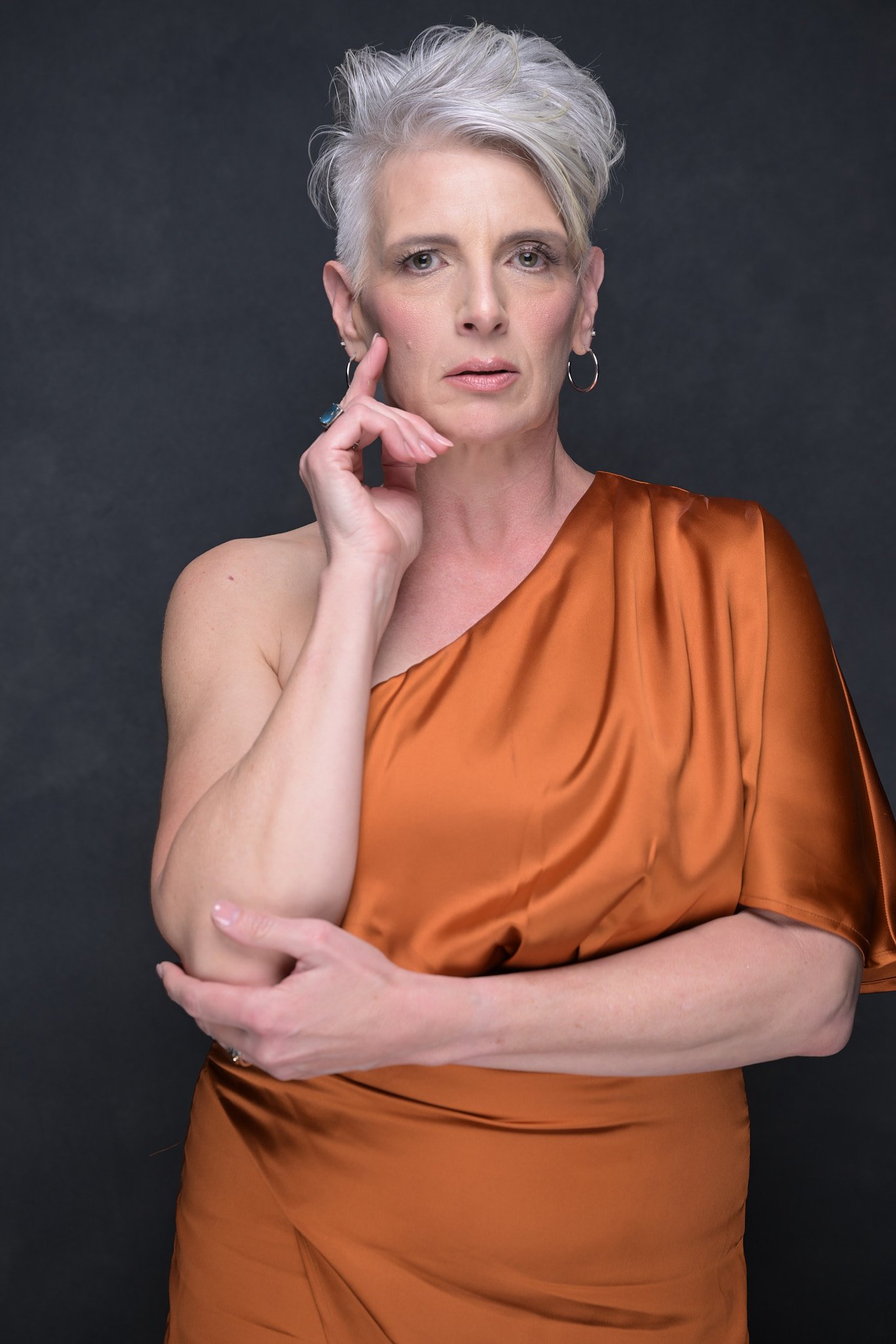 A woman with short gray hair, wearing a one-shoulder rust-colored satin dress, poses with her hand touching her face against a dark gray background.   Empowering women, headshots and branding, age campaign