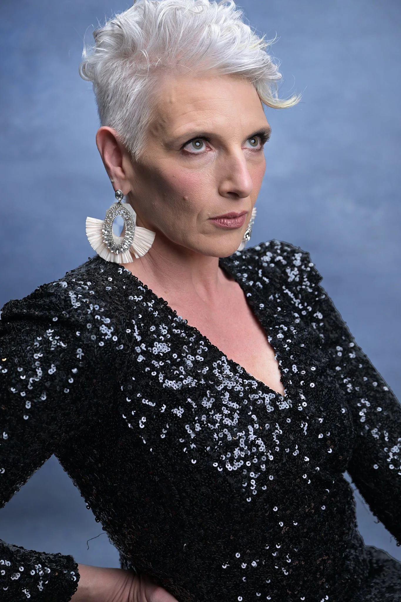 A woman with short, platinum blonde hair styled upward, wearing large, circular earrings with a ruffled design, and a black, sequined top with a v-neckline, posing against a cloudy blue sky background.   Empowering women, headshots and branding, age 