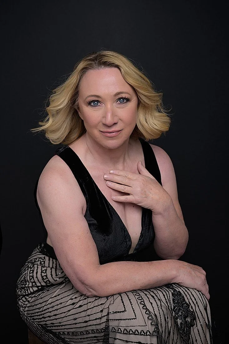 A woman with blonde, wavy hair poses against a black background, wearing a black sleeveless top and patterned skirt, with her hand resting on her chest and a confident expression.   Empowering women, headshots and branding, age campaign