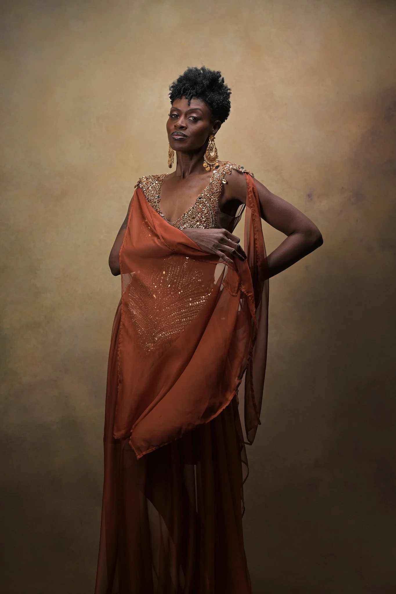A woman with short curly hair wearing an ornate, flowing, brown dress with gold embellishments and large gold earrings, standing against a neutral background.