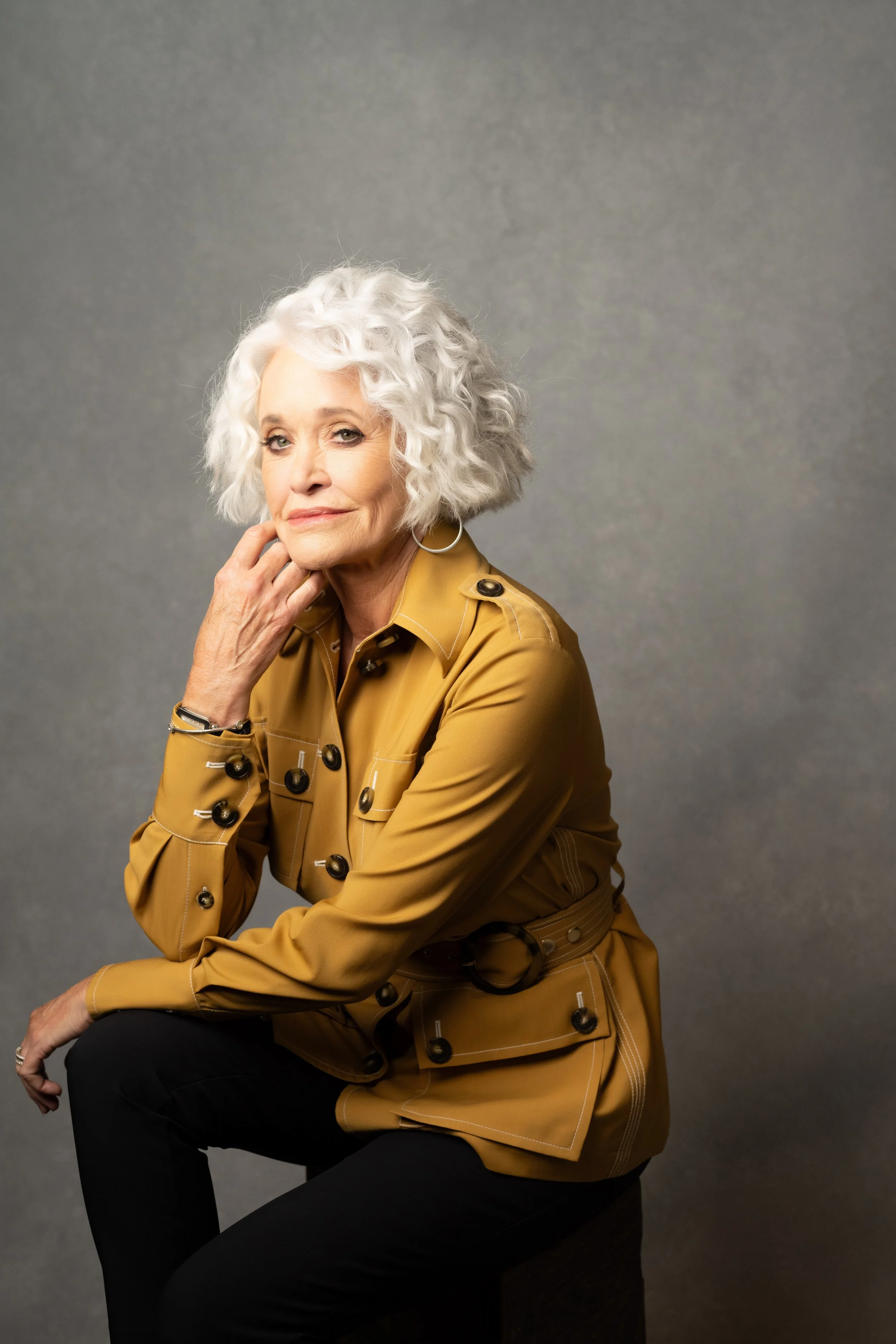 Senior woman with curly white hair wearing a tan trench coat and black pants, sitting against a plain gray background.   Empowering women, headshots and branding, age campaign