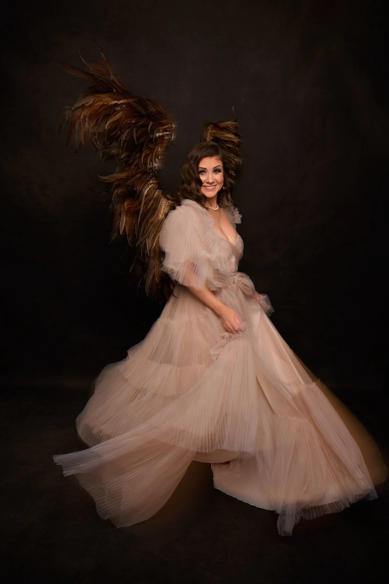 Woman wearing a beige tulle dress with large feathered wings against a dark background.   Empowering women, headshots and branding, age campaign