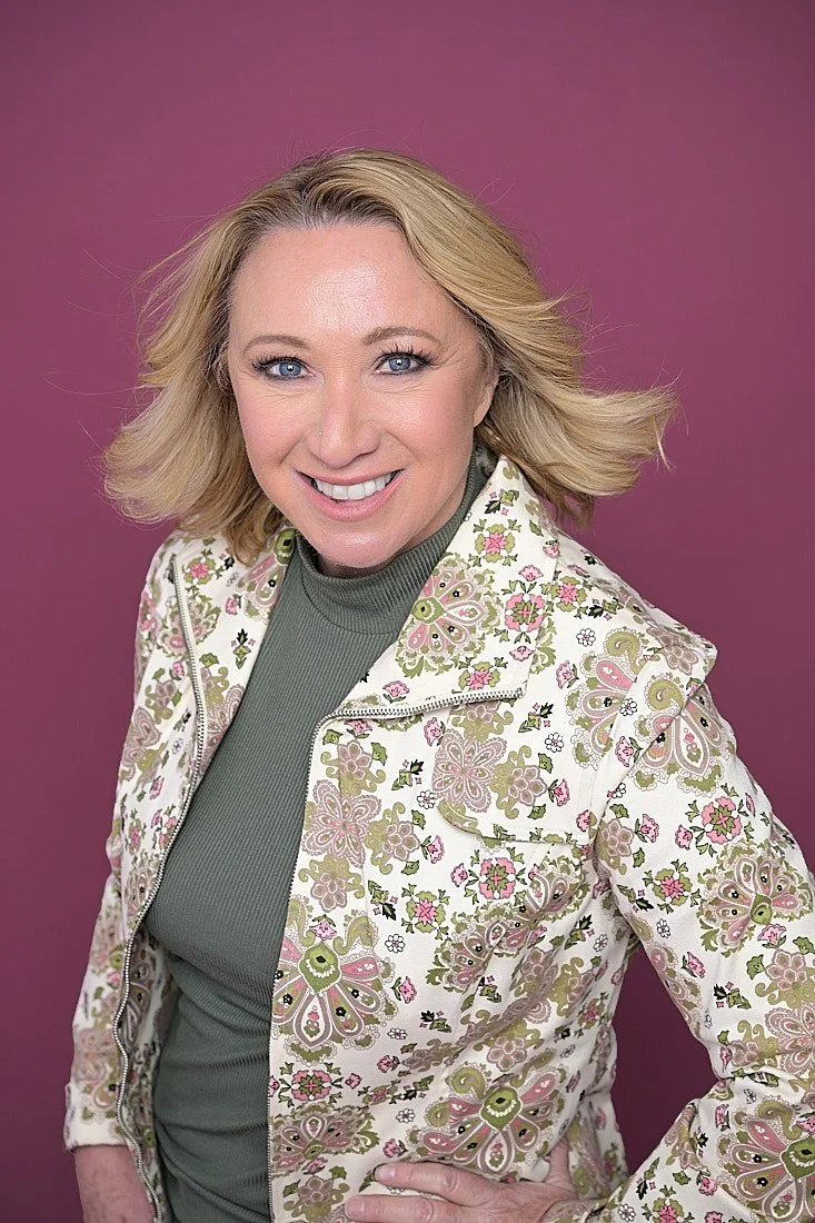 A woman with blonde hair wearing a floral-patterned jacket over a green turtleneck, smiling at the camera against a purple background.   Empowering women, headshots and branding, age campaign