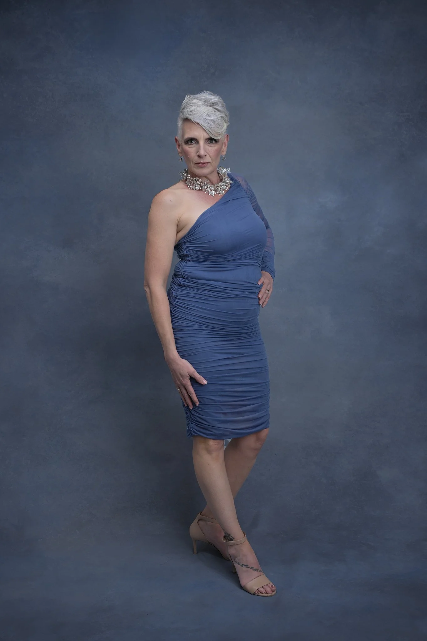 A woman with short, silver hair wearing a form-fitting, blue, ruched dress and beige high heels poses against a grey textured background.   Empowering women, headshots and branding, age campaign