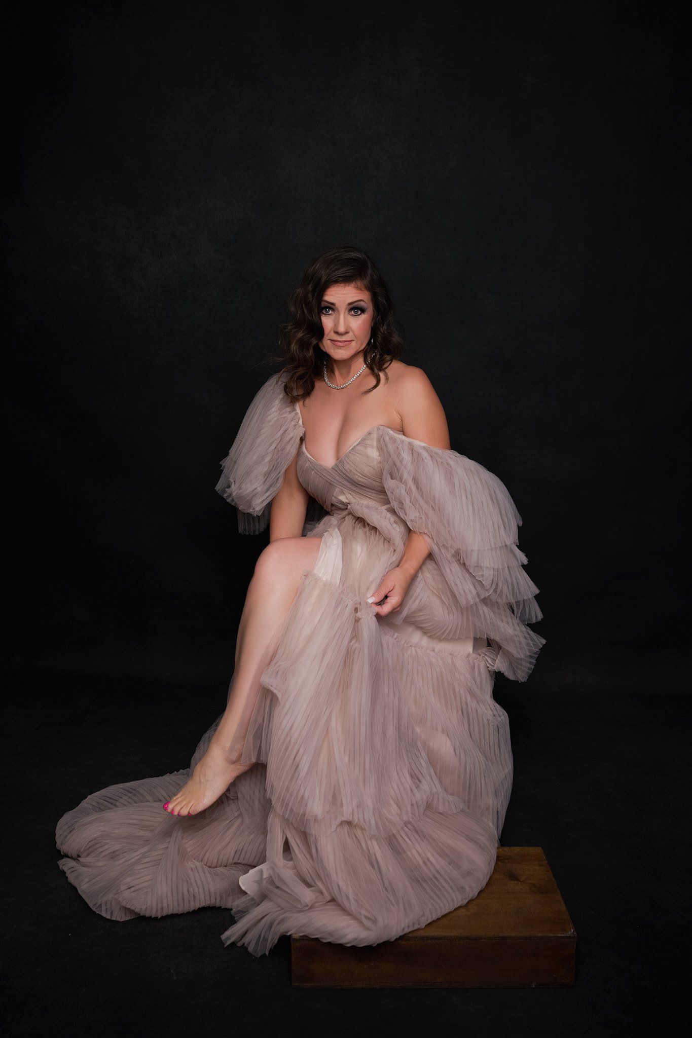 A woman in a beige, flowing, ruffled gown sitting on a wooden block against a black backdrop, looking at the camera.   Empowering women, headshots and branding, age campaign