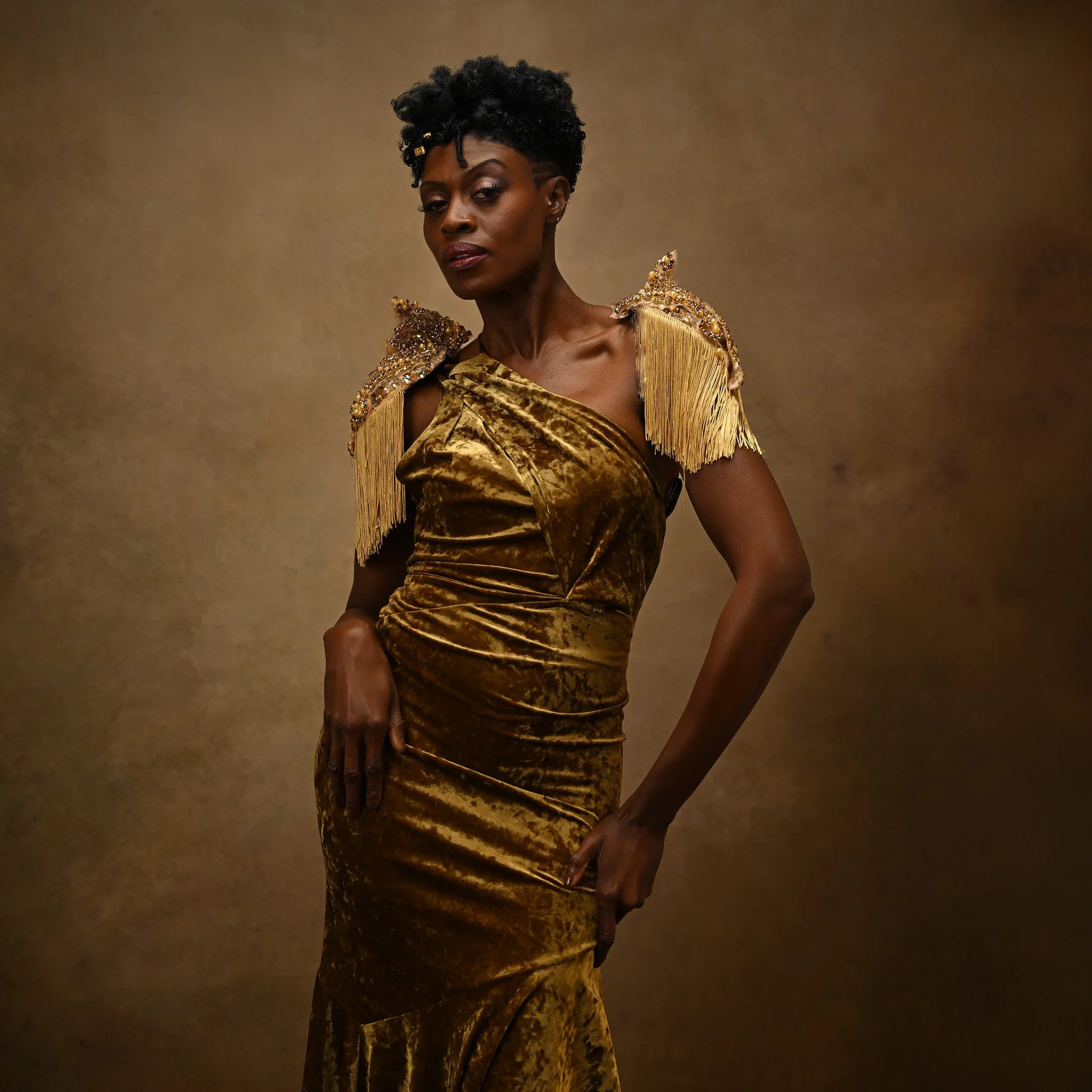 A woman in a gold velvet gown with ornate shoulder embellishments and fringe details, standing against a brown background.   Empowering women, headshots and branding, age campaign