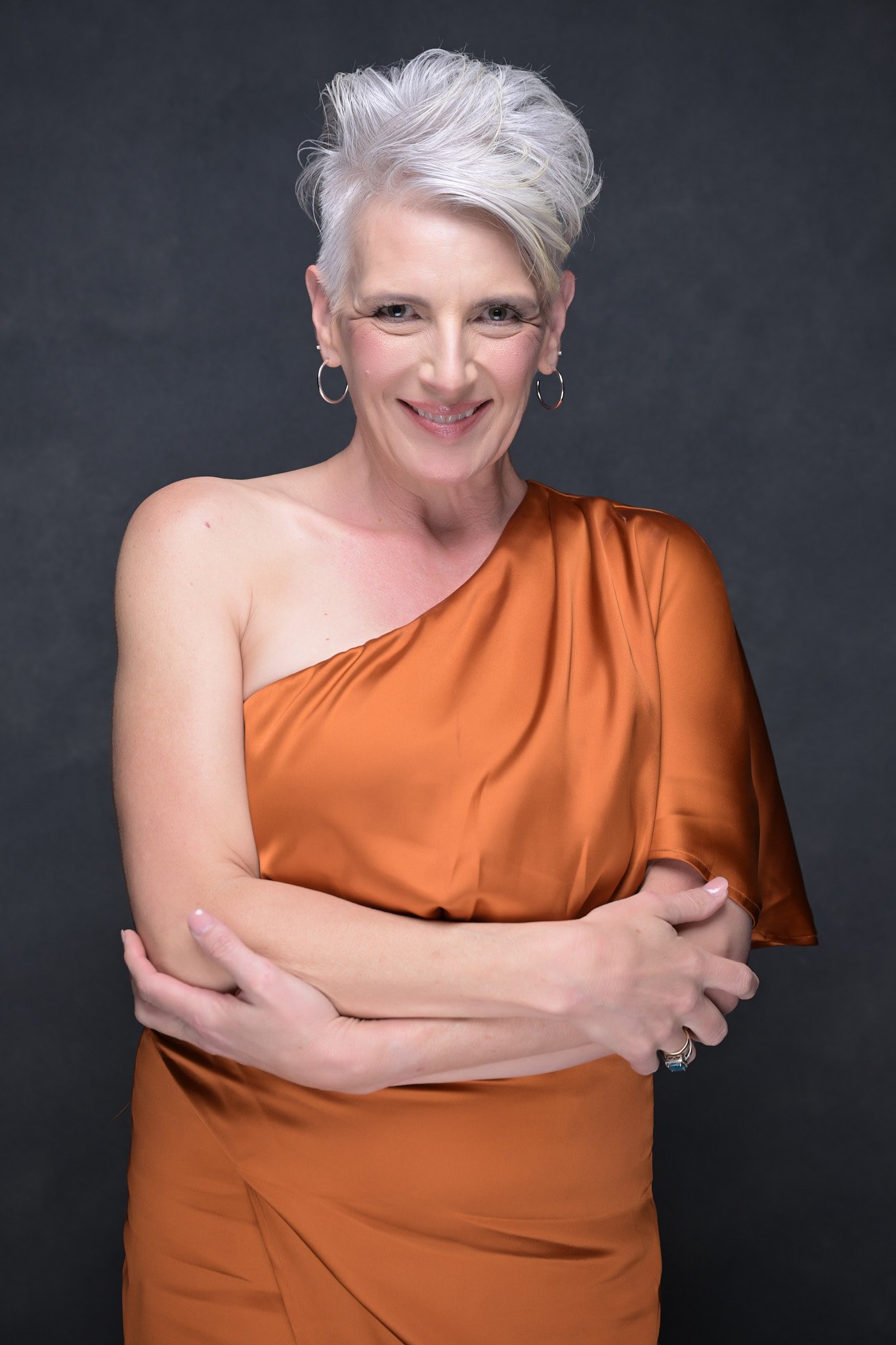 Portrait of a smiling mature woman with short gray hair, wearing an orange satin dress and hoop earrings, against a dark background.   Empowering women, headshots and branding, age campaign