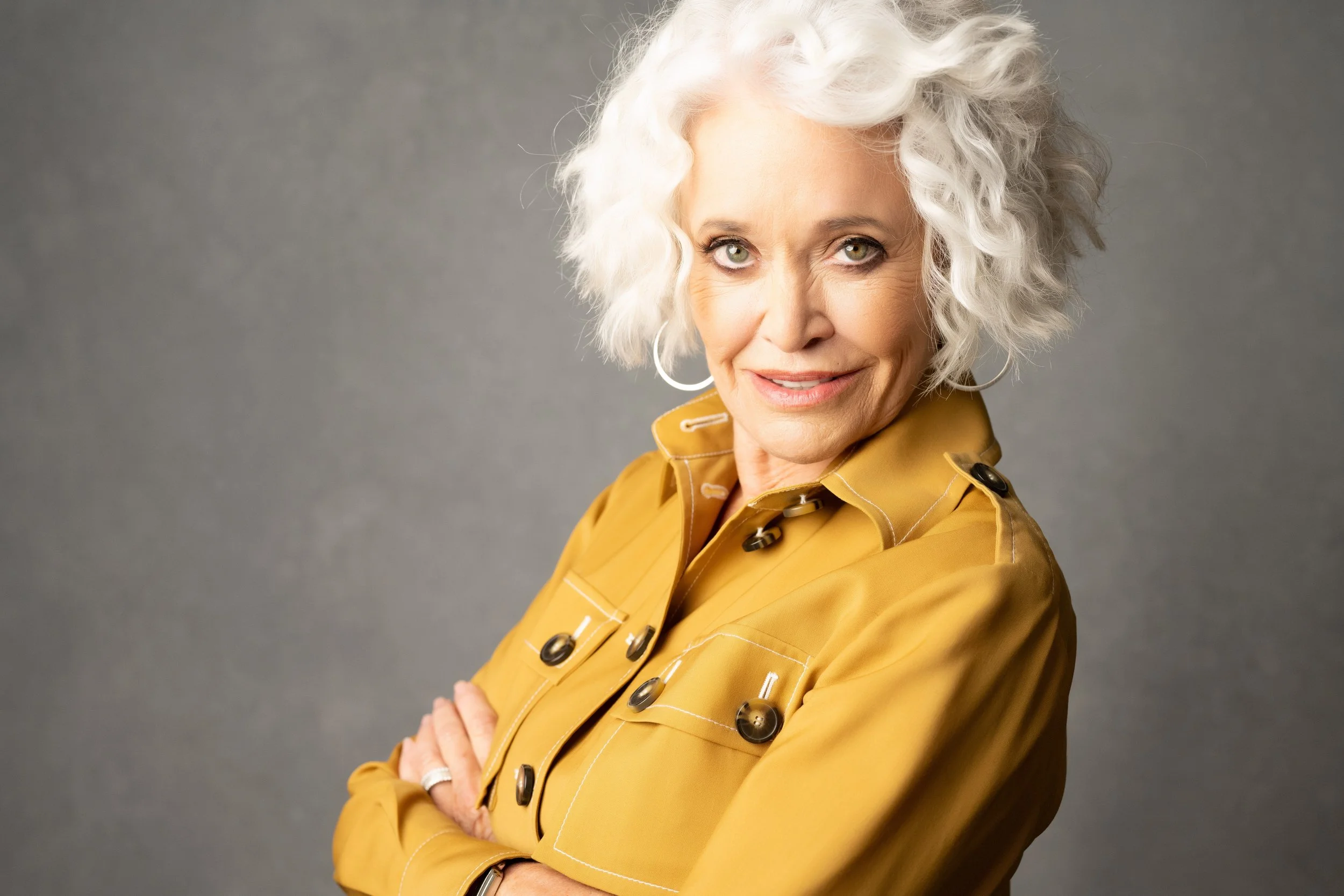 An older woman with curly white hair smiling, wearing a mustard yellow jacket with black buttons and hoop earrings, standing against a gray background.  Empowering women, headshots and branding, age campaign