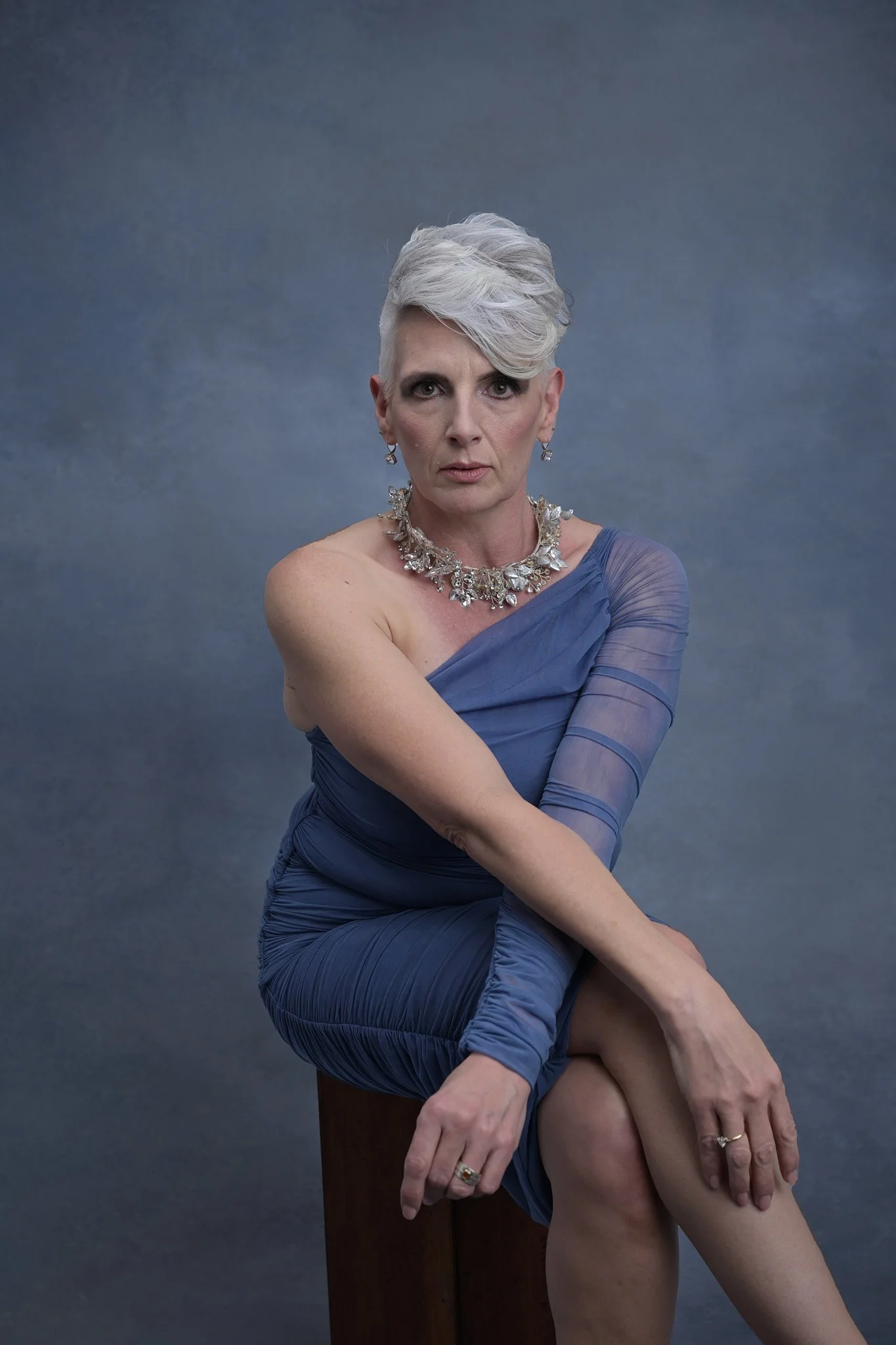 An older woman with short, white hair styled to the side, wearing a one-shoulder blue dress with sheer sleeve details, a large statement necklace, and earrings, sitting on a wooden block in front of a plain gray background, looking directly at the ca