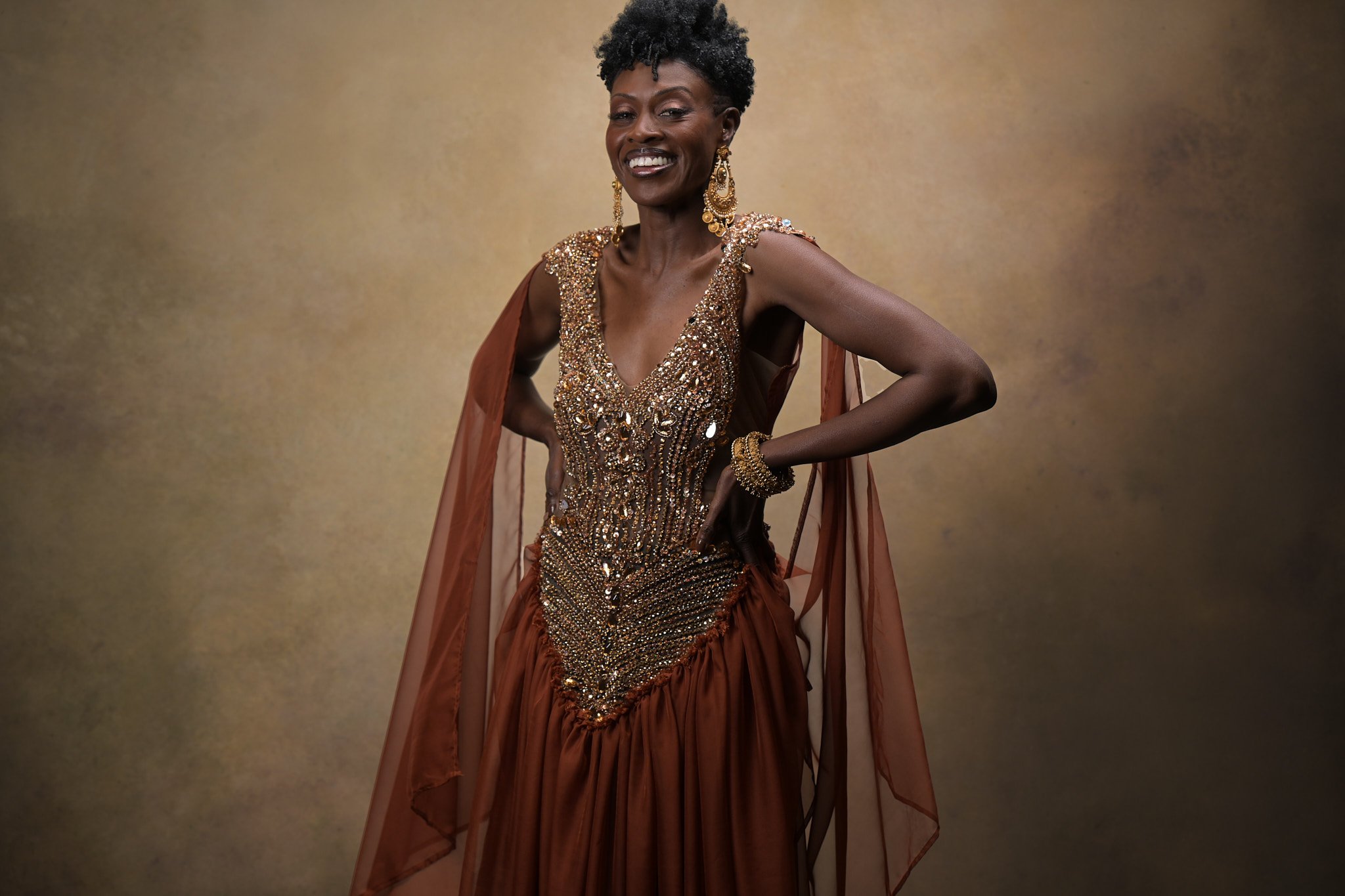 Elegant woman in a shimmering gold and brown gown with ornate earrings and bracelets, posing confidently against a gold-toned background. Empowering women, headshots, and Branding sessions.  Age campaign.