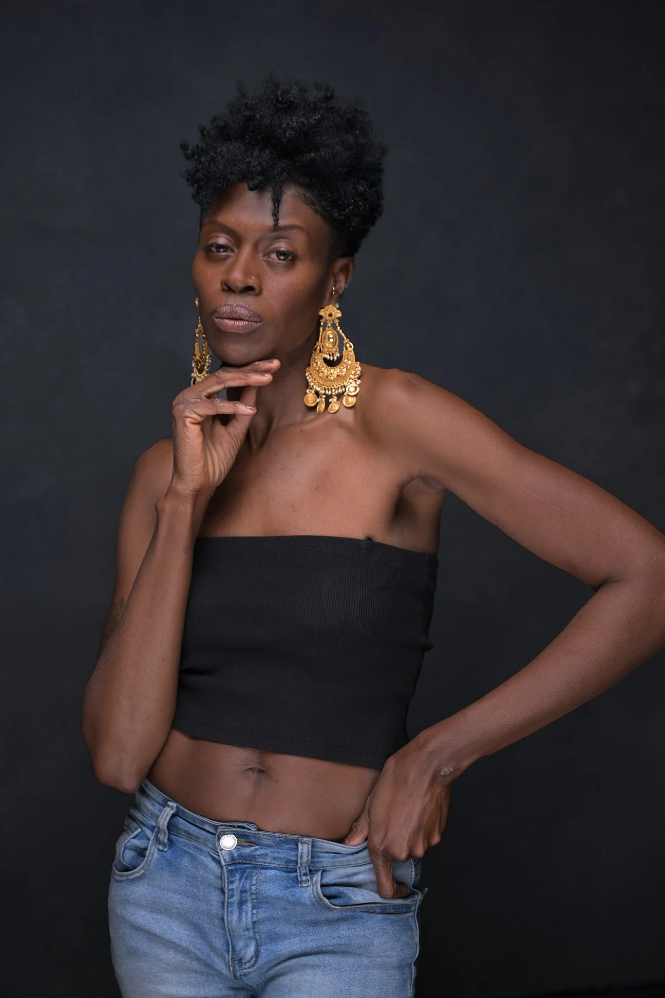 A woman with short, curly black hair wearing large gold earrings, a black strapless top, and blue jeans against a dark background. Empowering women, headshots, and Branding sessions.  Age campaign.