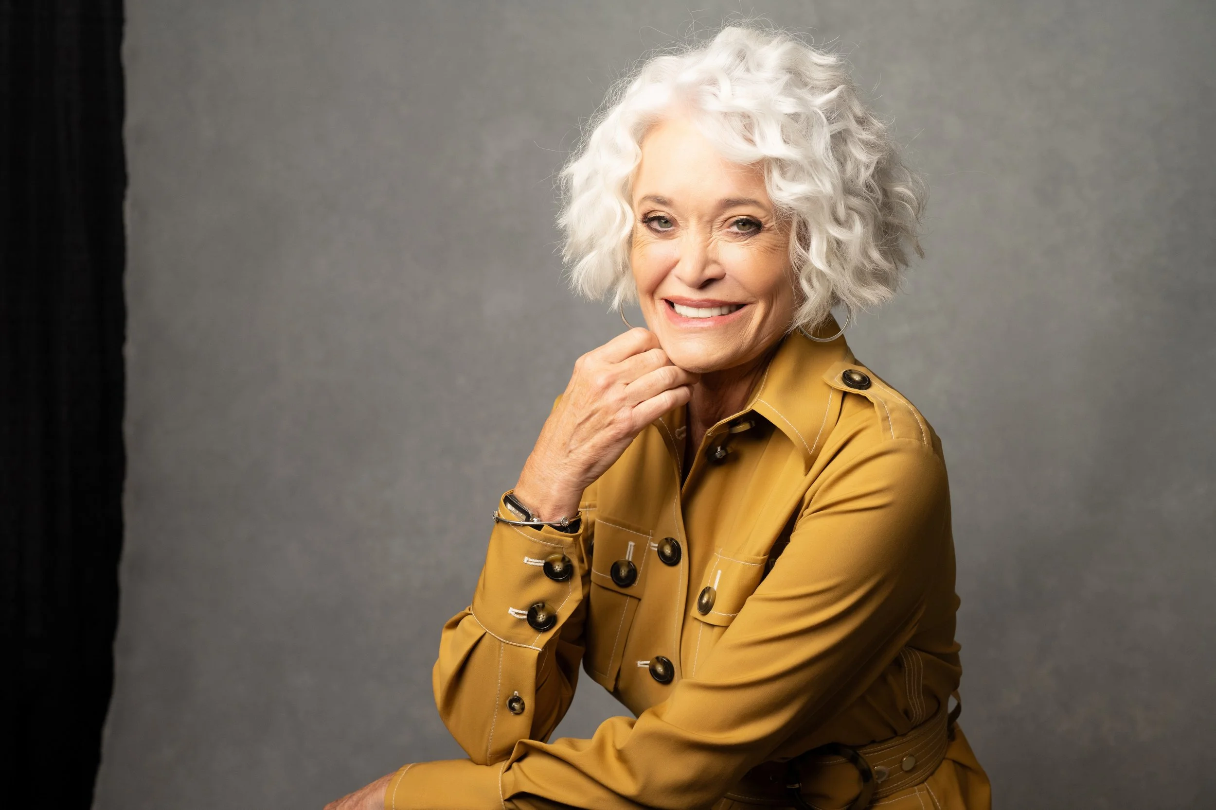 A smiling elderly woman with white curly hair, wearing a tan button-up jacket, sitting against a plain gray background.   Empowering women, headshots and branding, age campaign