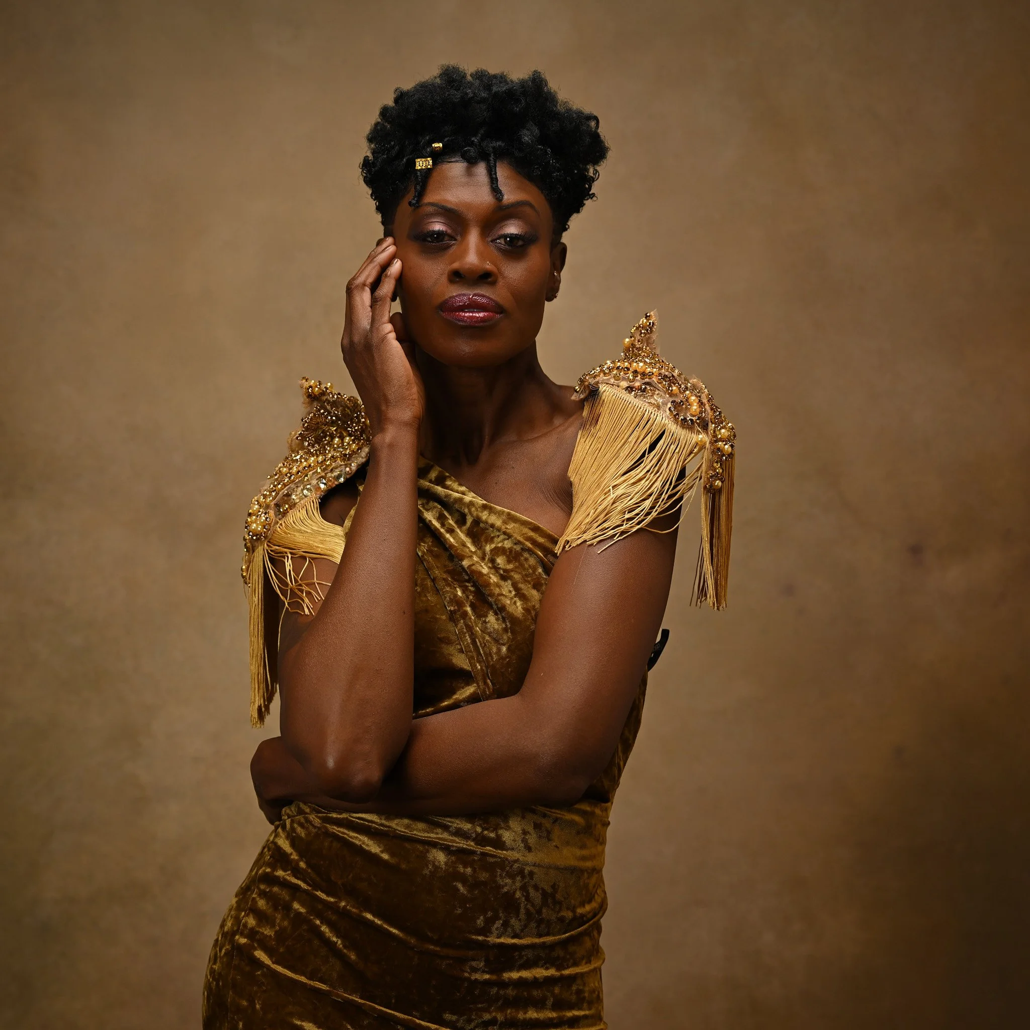 Elegant woman dressed in a gold velvet gown with ornate shoulder embellishments, posing with her hand near her face against a gold background.   Empowering women, headshots and branding, age campaign