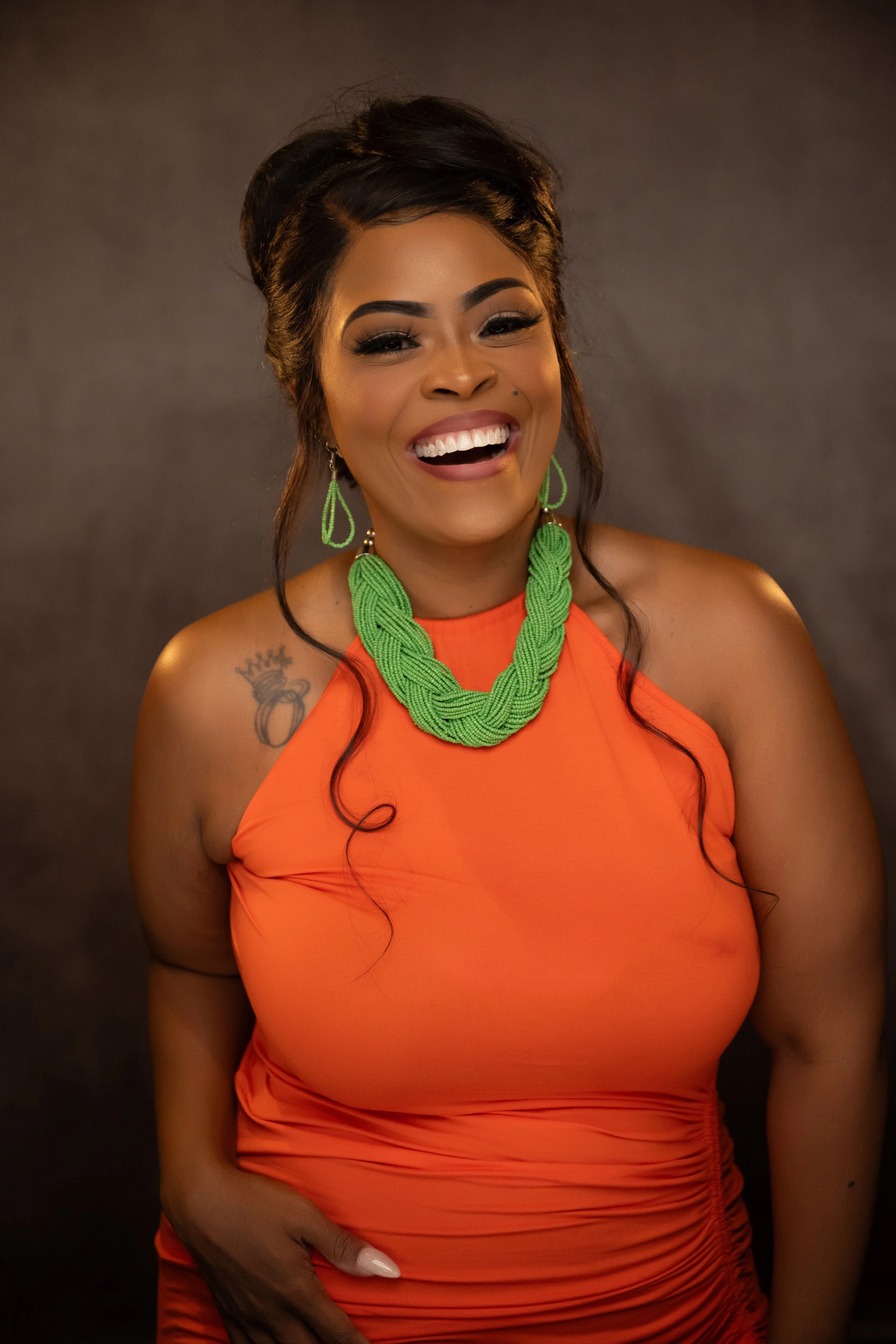 A smiling woman with dark hair styled in waves, wearing an orange sleeveless dress, green beaded jewelry, and a tattoo of a crown and ring on her shoulder, standing against a dark background.   Empowering women, headshots and branding, age campaign