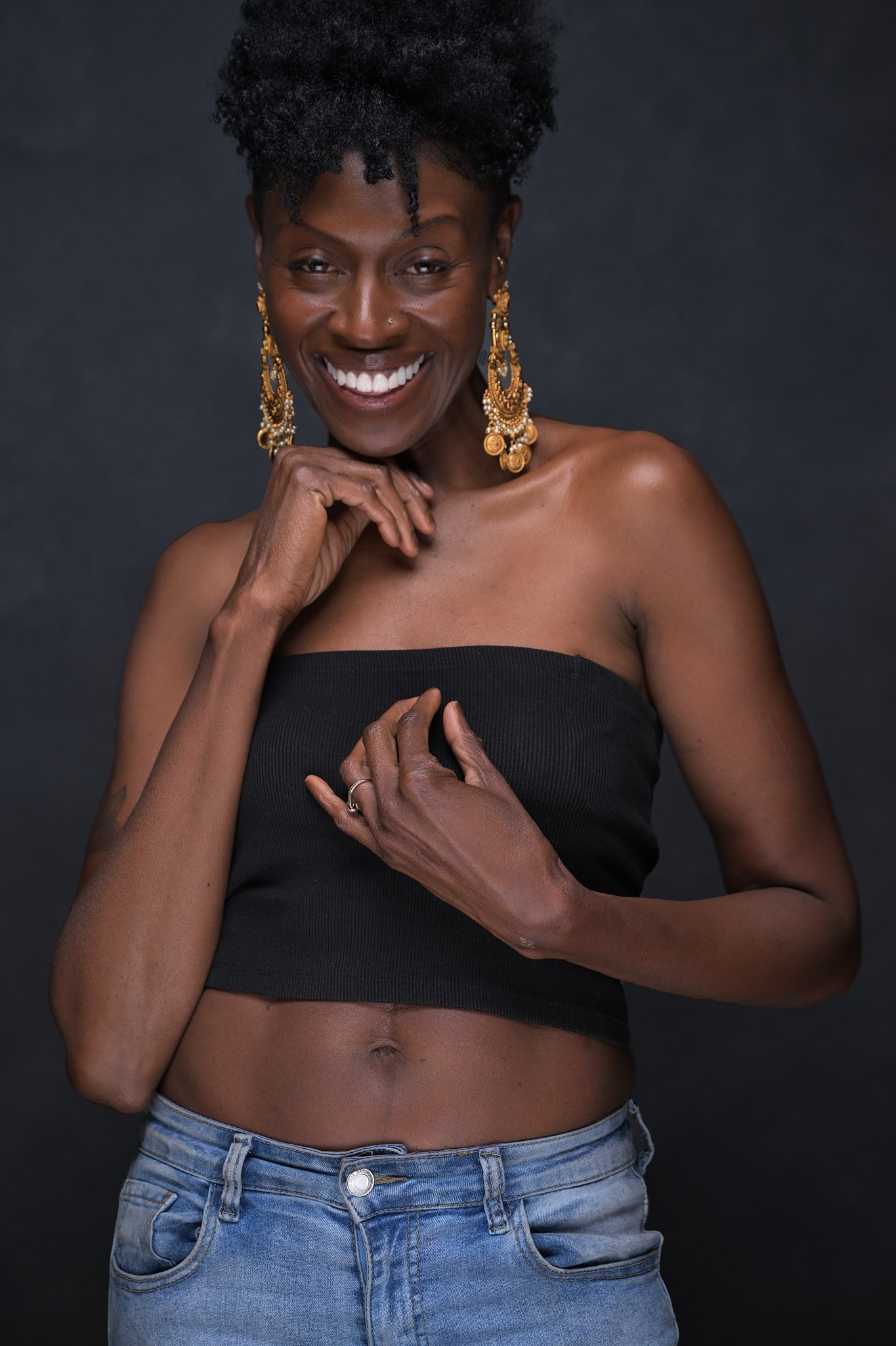 Portrait of a smiling woman with dark skin, wearing gold earrings, a black tube top, and blue jeans, against a dark background. Empowering women, headshots, and Branding sessions.  Age campaign.
