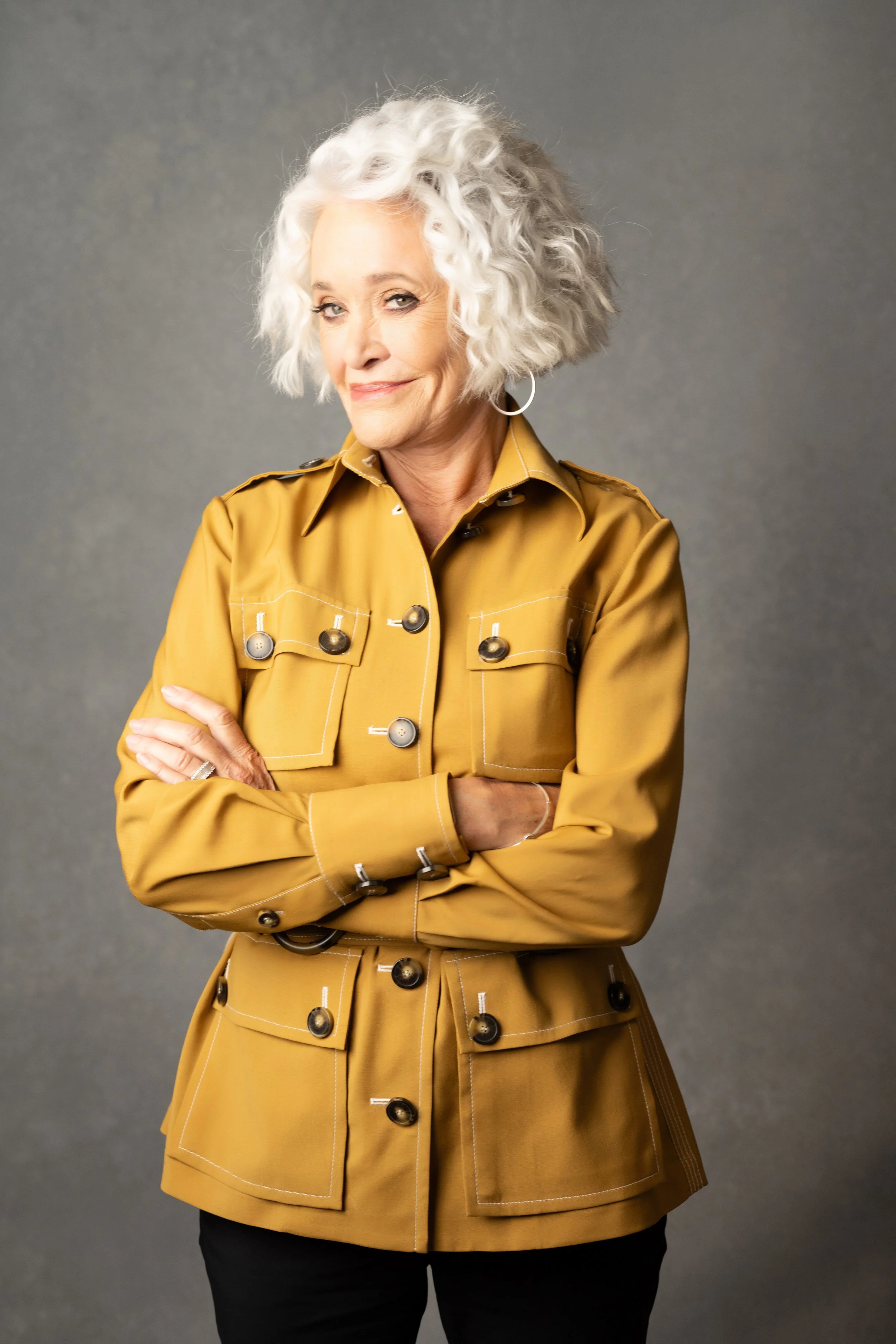 An older woman with curly white hair wearing a mustard yellow jacket with multiple pockets and buttons, standing with arms crossed against a neutral gray background.  Empowering women, headshots and branding