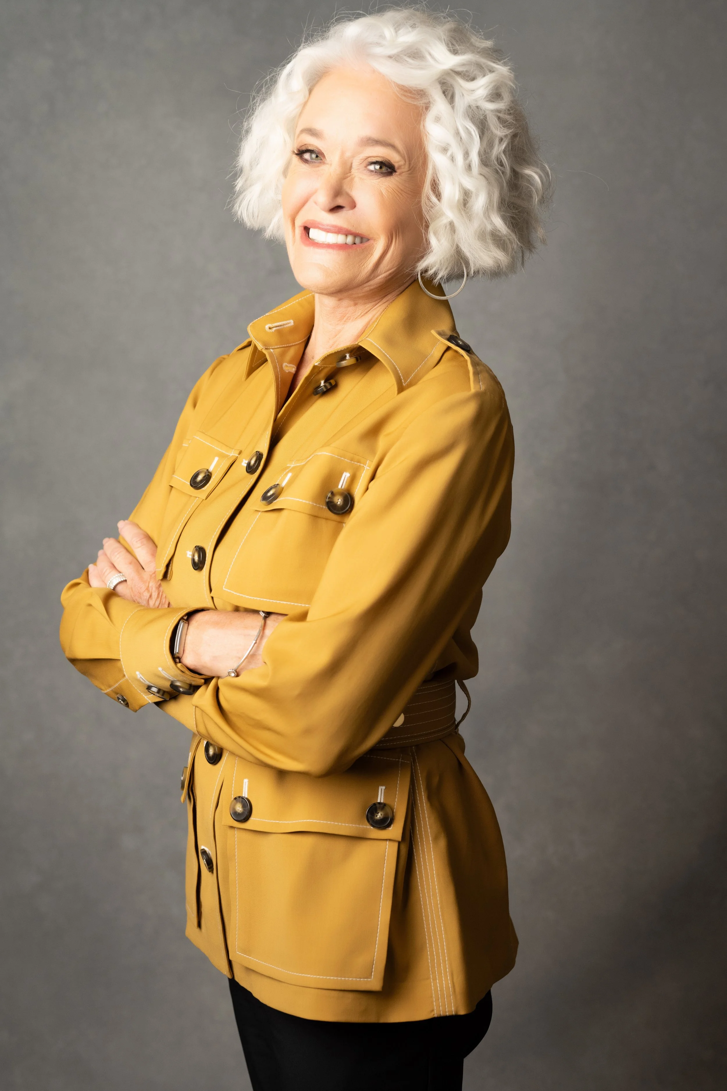 A cheerful elderly woman with curly white hair, wearing a mustard yellow jacket with multiple pockets, standing with arms crossed against a gray background.   Empowering women, headshots and branding, age campaign