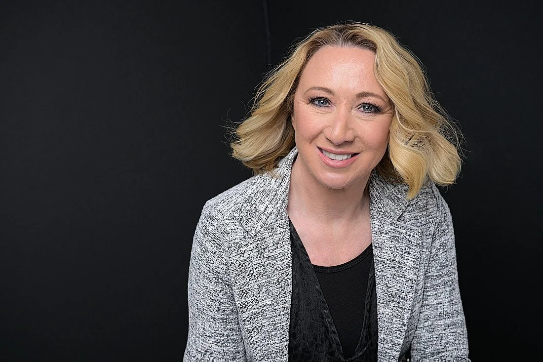 Headshot of a smiling woman with blonde hair, wearing a gray blazer over a black top, against a dark background.   Empowering women, headshots and branding, age campaign