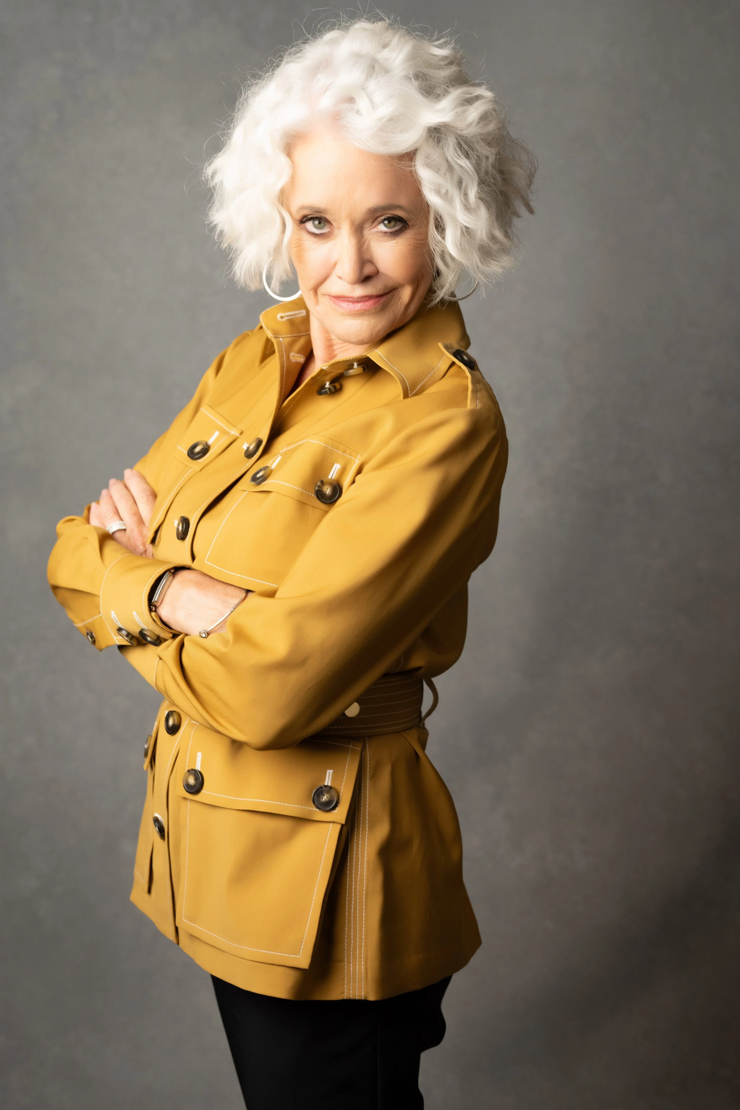 An older woman with curly white hair, wearing a mustard yellow jacket with black buttons, crosses her arms and looks directly at the camera with a slight smile. Empowering women, headshots and branding