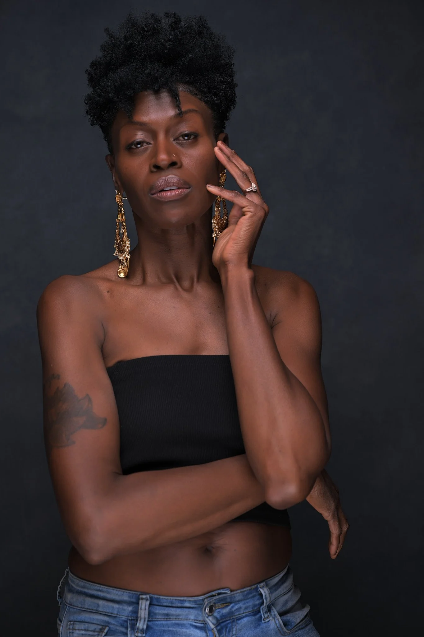 A confident woman with natural curly hair, wearing a black strapless top, blue jeans, and ornate gold earrings, standing against a dark background. Empowering women, headshots, and Branding sessions.  Age campaign.