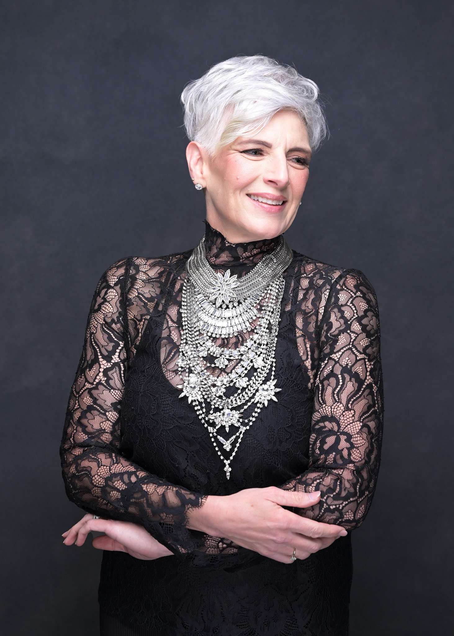 A woman with short silver hair wearing a black lace dress and multiple layered silver necklaces, standing against a dark background.   Empowering women, headshots and branding, age campaign