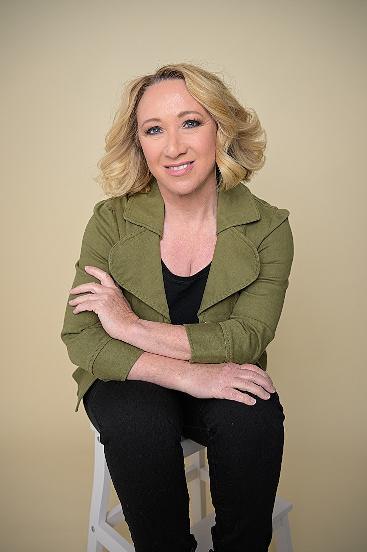 A woman with blonde, curly hair sitting on a stool, wearing a green jacket over a black top, with a light beige background.   Empowering women, headshots and branding, age campaign