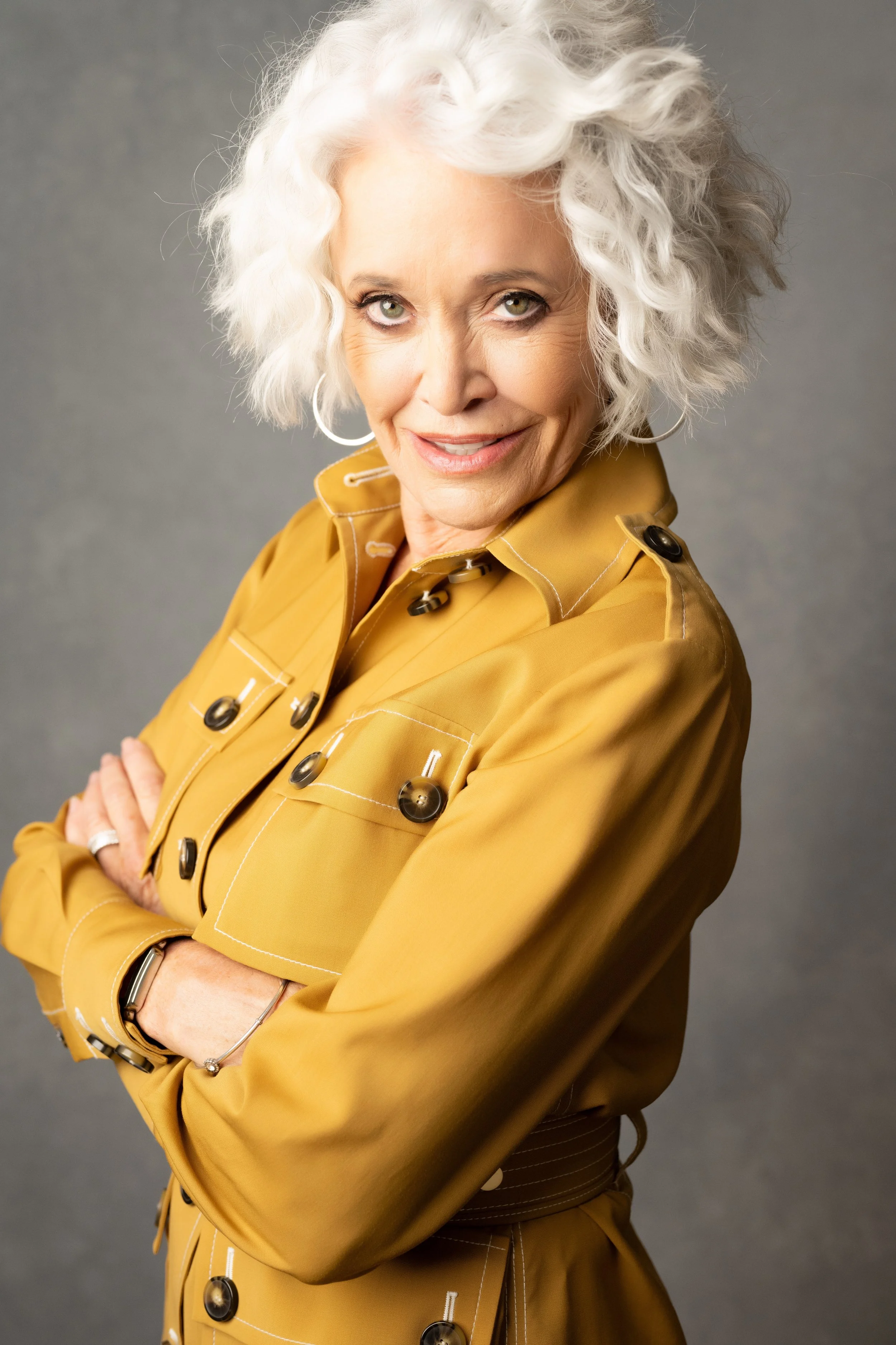 An older woman with curly white hair, wearing a mustard-colored trench coat, standing with arms crossed and smiling slightly.  Empowering women, headshots and branding, age campaign