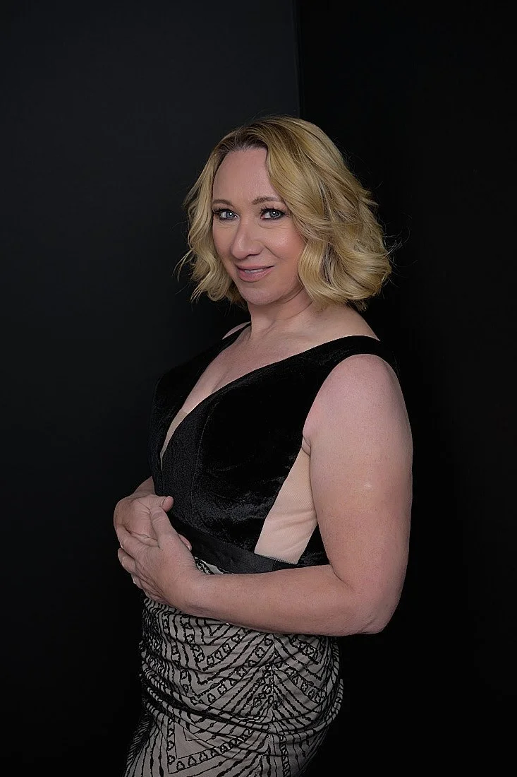 A woman with blonde, wavy hair wearing a sleeveless black and beige dress, standing against a dark background and looking at the camera.   Empowering women, headshots and branding, age campaign