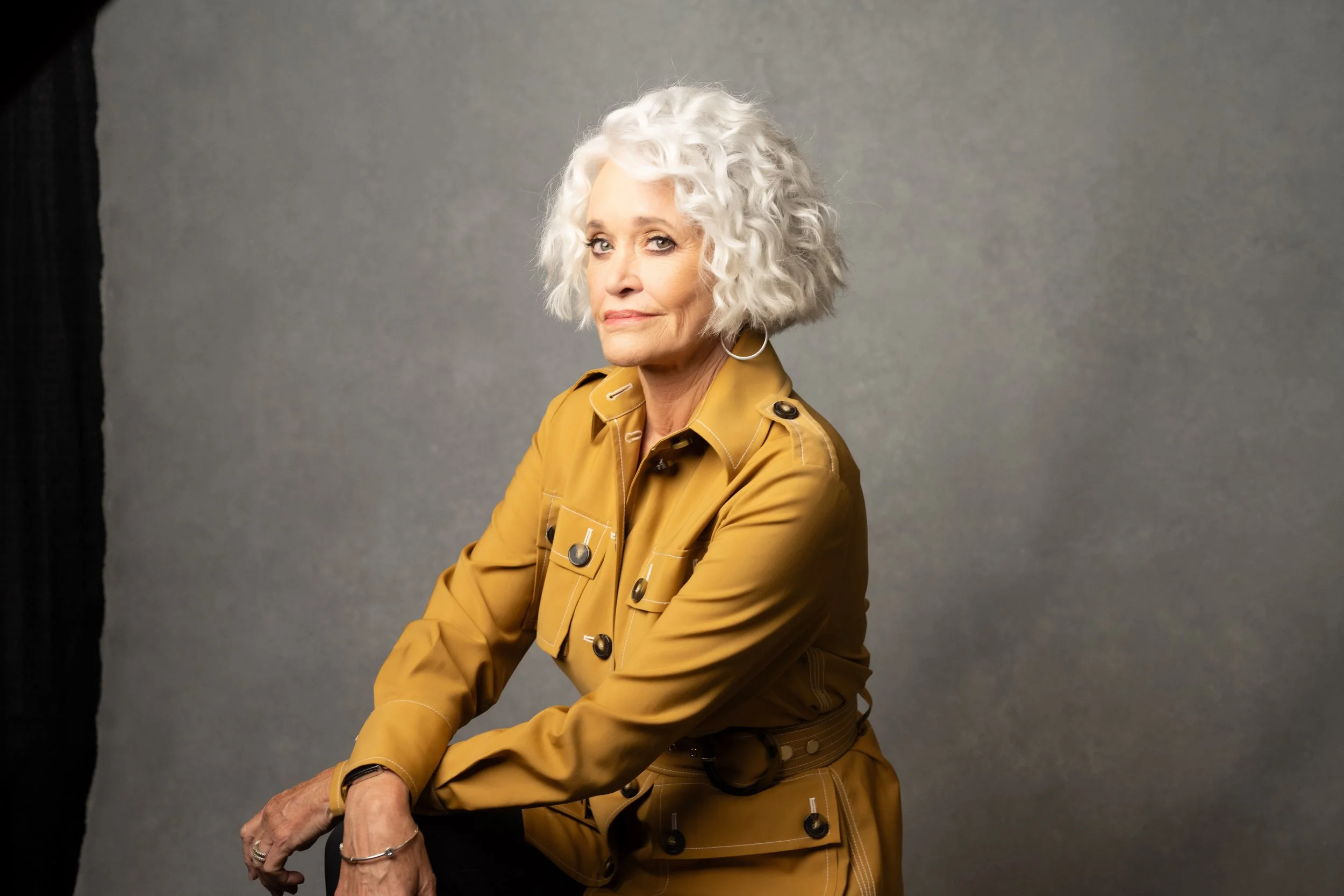 An older woman with curly white hair, wearing a yellow jacket with black buttons, sitting against a plain gray background.   Empowering women, headshots and branding, age campaign