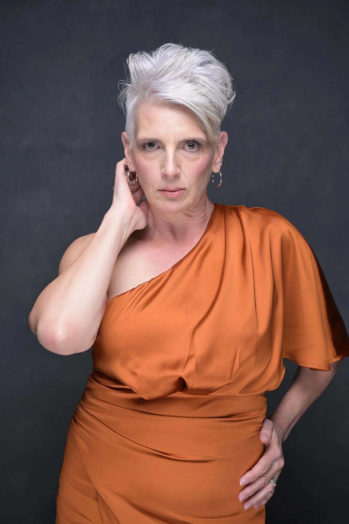 Woman with short, white hair, wearing a one-shoulder, bronze satin dress against a dark background.   Empowering women, headshots and branding, age campaign