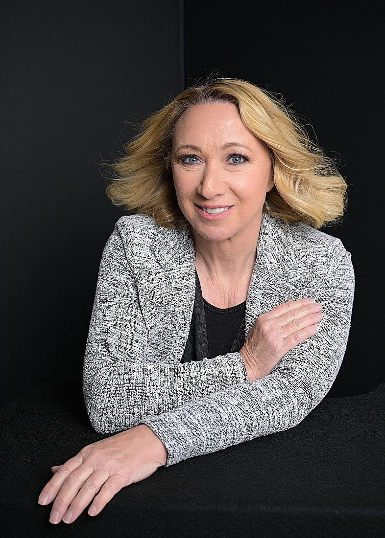 A smiling blonde woman in a gray blazer, sitting at a black table against a black background.   Empowering women, headshots and branding, age campaign