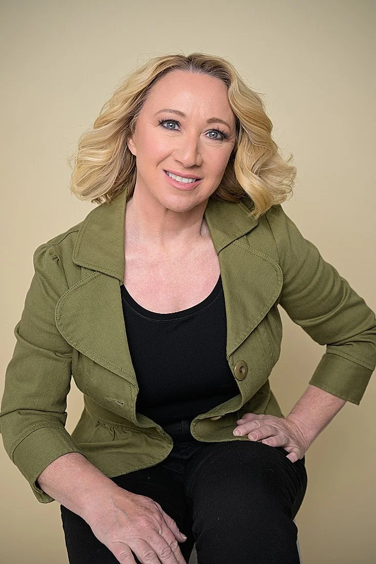 A woman with blonde, wavy hair, wearing a green jacket over a black top, sitting against a beige background and smiling at the camera.   Empowering women, headshots and branding, age campaign