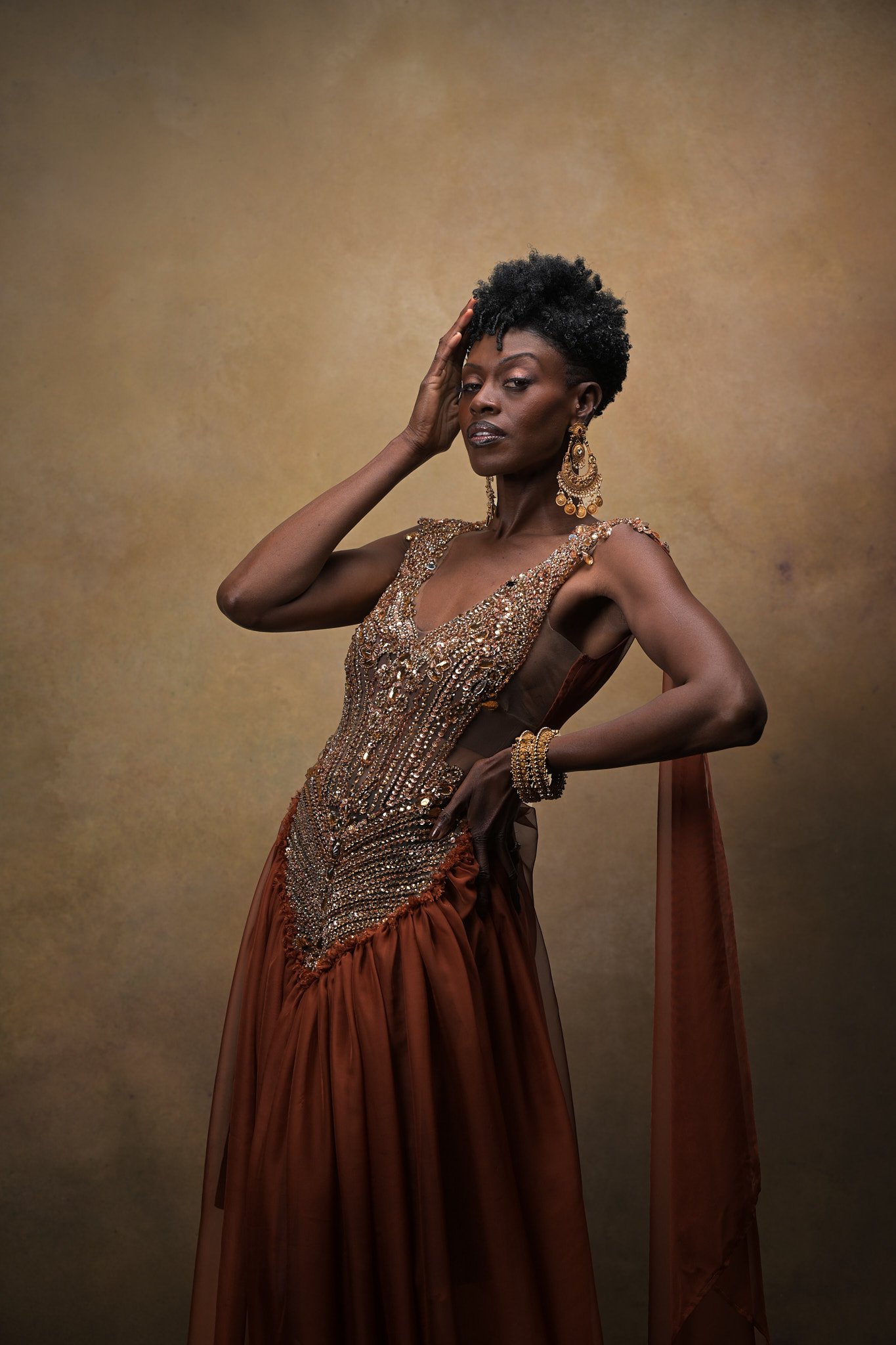 A woman with short curly black hair and dark skin tone dressed in an ornate, jewel-toned gown with gold embellishments and jewelry, posing against a plain brownish backdrop.   Empowering women, headshots and branding, age campaign