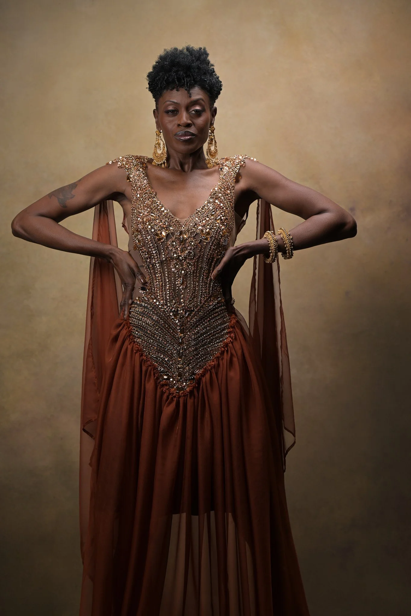Woman in an ornate, beaded brown gown with sheer cape, wearing large gold earrings and bracelets, standing confidently with hands on hips. Empowering women, headshots, and Branding sessions.  Age campaign.