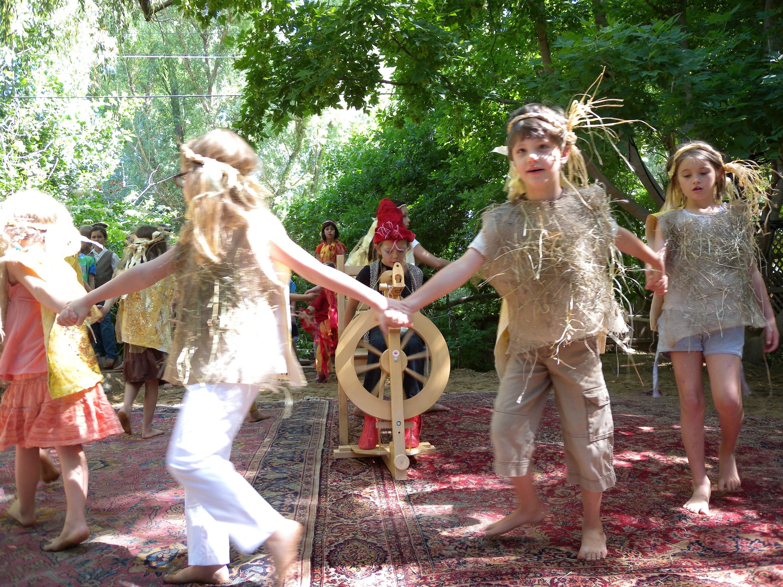 During a theater performance of the Rumplestiltskin Play at Sage Programs in Boulder CO, children dressed in handmade costumes with straw and natural materials, holding hands in a circle outdoors on a patterned rug.
