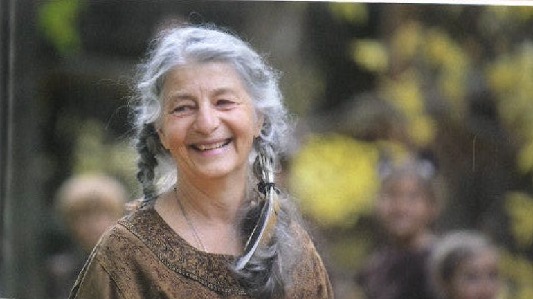 Sage Hamilton, a joyful elderly woman with gray hair in braided pigtails, smiling outdoors in a natural setting during a theater performance at Sage Programs in Boulder, Colorado.