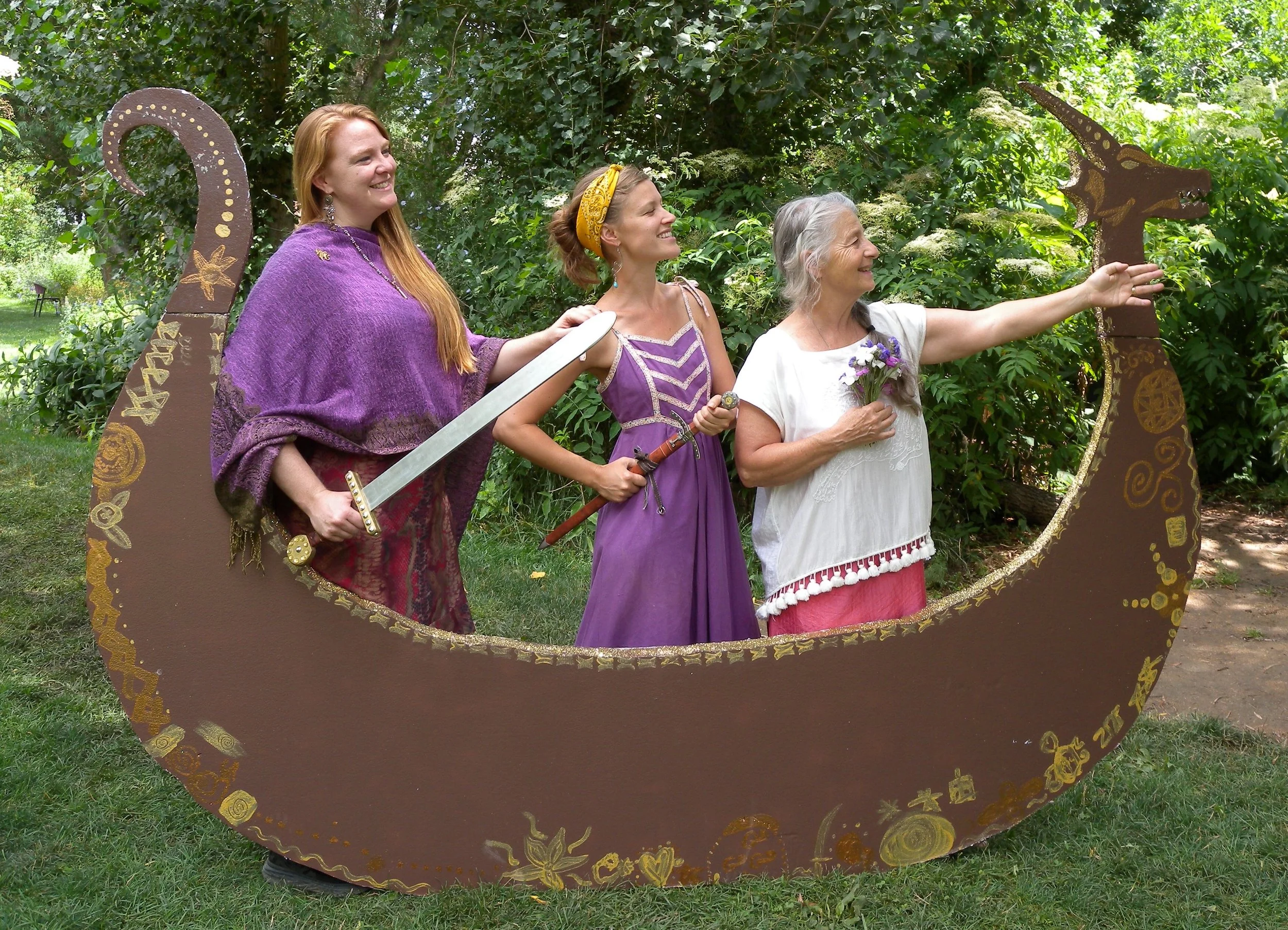 During a play at Sage Programs in Boulder Coloraod, three women dressed in fantasy-style costumes, holding swords and flowers, standing behind a large, crescent-shaped prop decorated with gold patterns and a toy dragon on top.