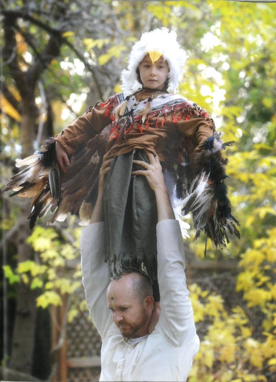 A man is holding a young girl dressed in a Native American costume with feathers, in an outdoor setting with trees and yellow leaves. This is part of the Jumping Mouse Play Performance at Sage Programs in Boulder CO.