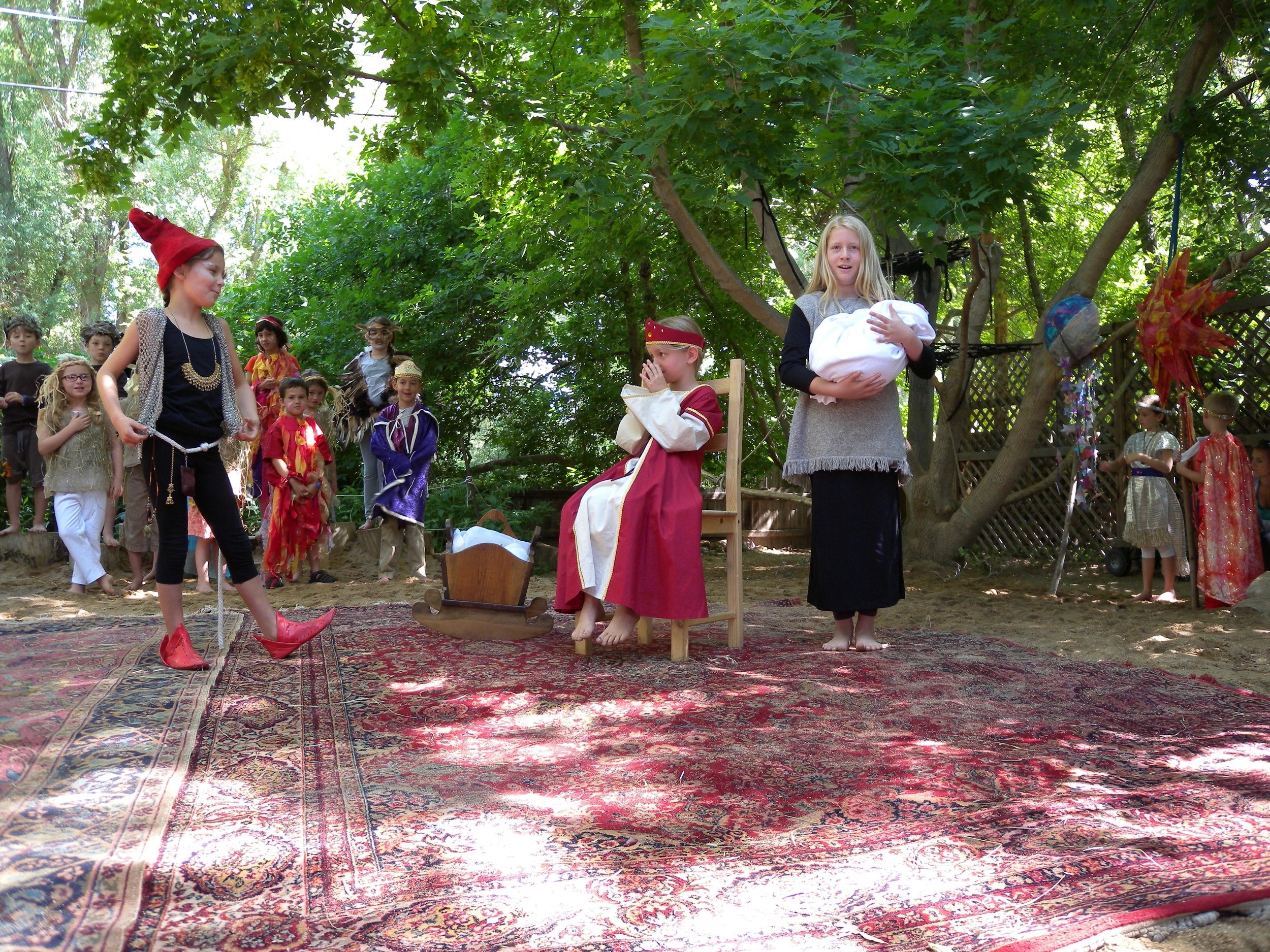 Children dressed as characters in a Nativity scene, with a girl holding a baby doll, a boy in a robe sitting on a chair, and others in costumes, outdoors under trees.