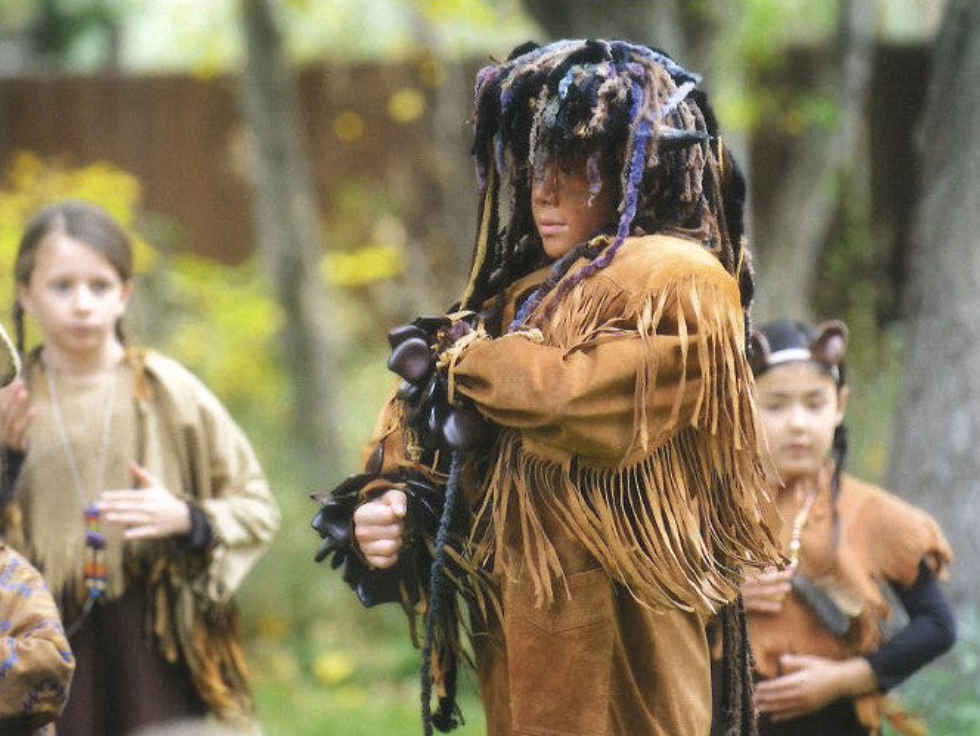 Children dressed as Native Americans with fringed clothing and accessories, outdoors in a wooded area. This is the performance of the Jumping Mouse Play at Sage Programs, in Boulder Colorado.