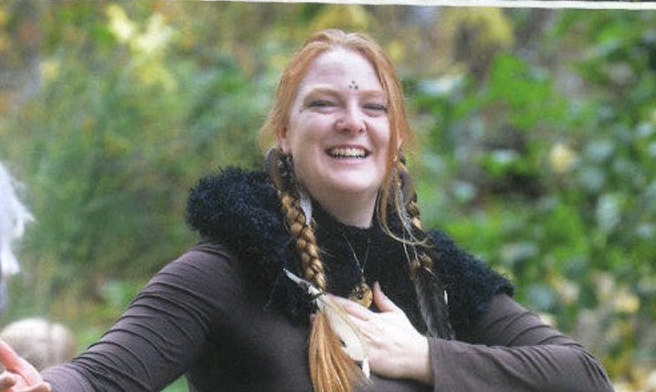 Meagan Chandler is smiling and has red hair, braided, wearing a dark outfit with a fur collar, standing outdoors during a theater performance at Sage Programs in Boulder, Colorado.