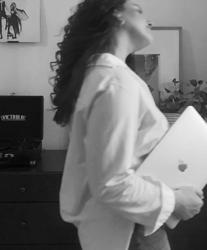 A woman with curly hair wearing a light-colored shirt holding a laptop in a room with a dresser, a record player, a framed picture, and a plant.