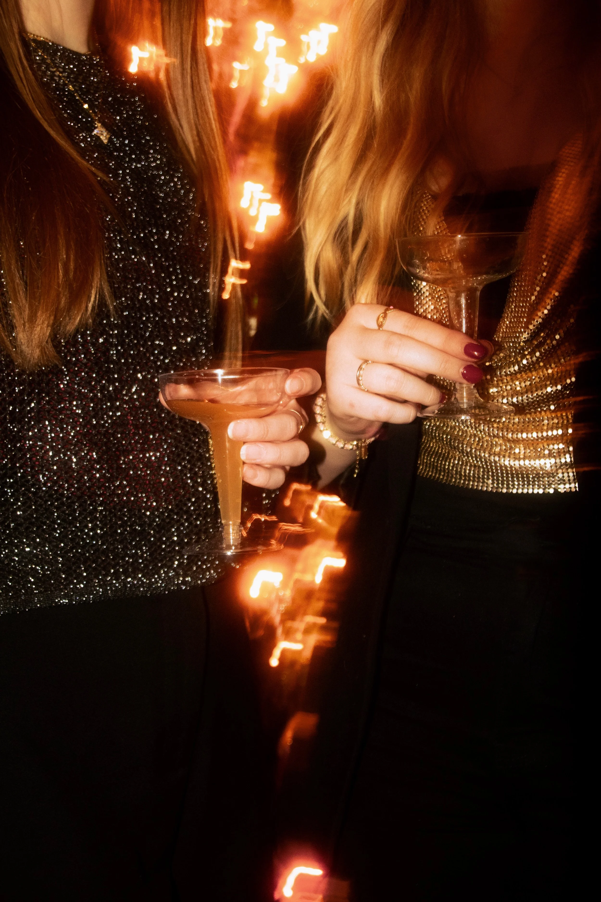 Two women in glittery dresses holding cocktail glasses at a celebration with blurred festive lights in the background.